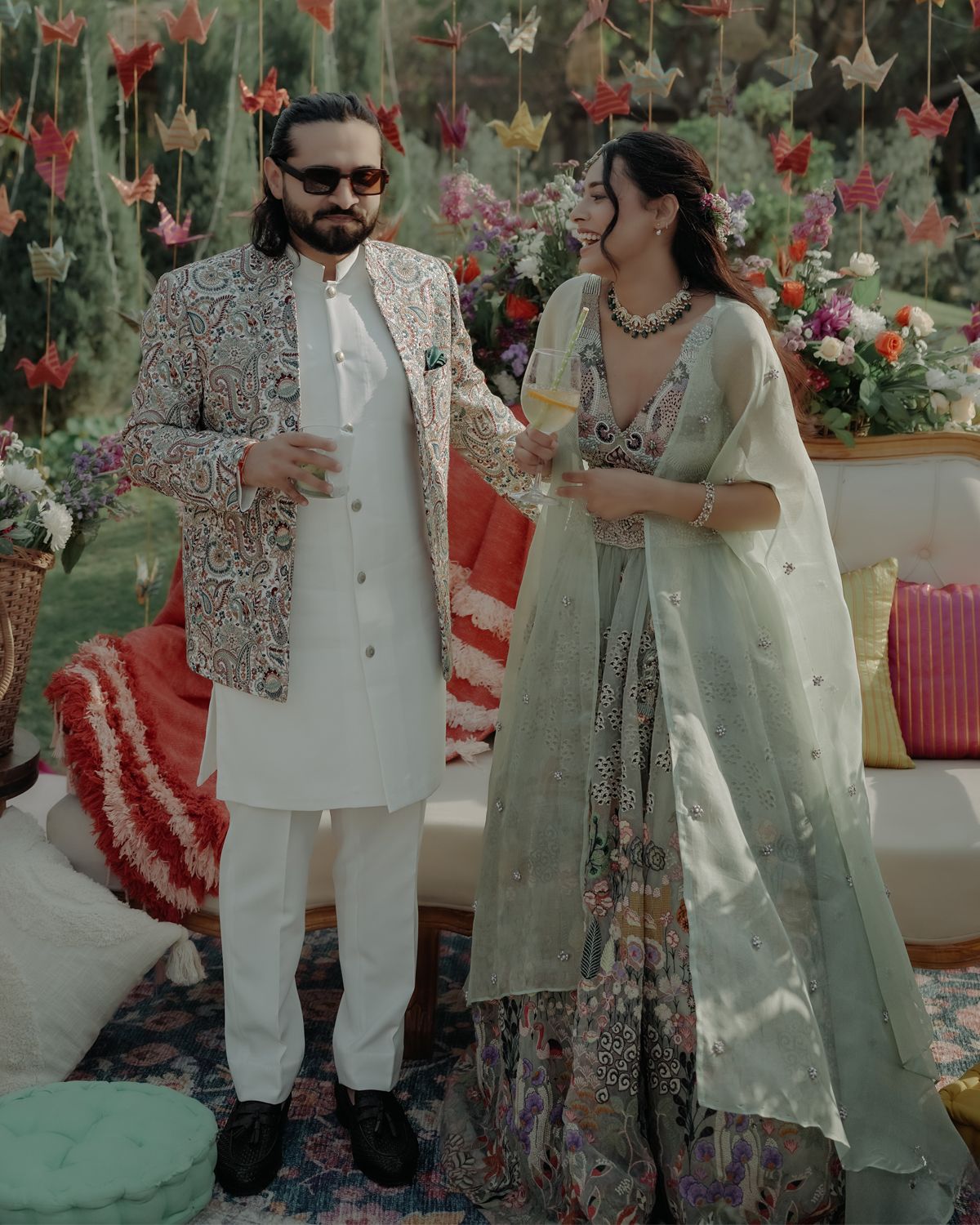 A couple in traditional South Asian attire share a joyful moment at an outdoor celebration with floral decorations.