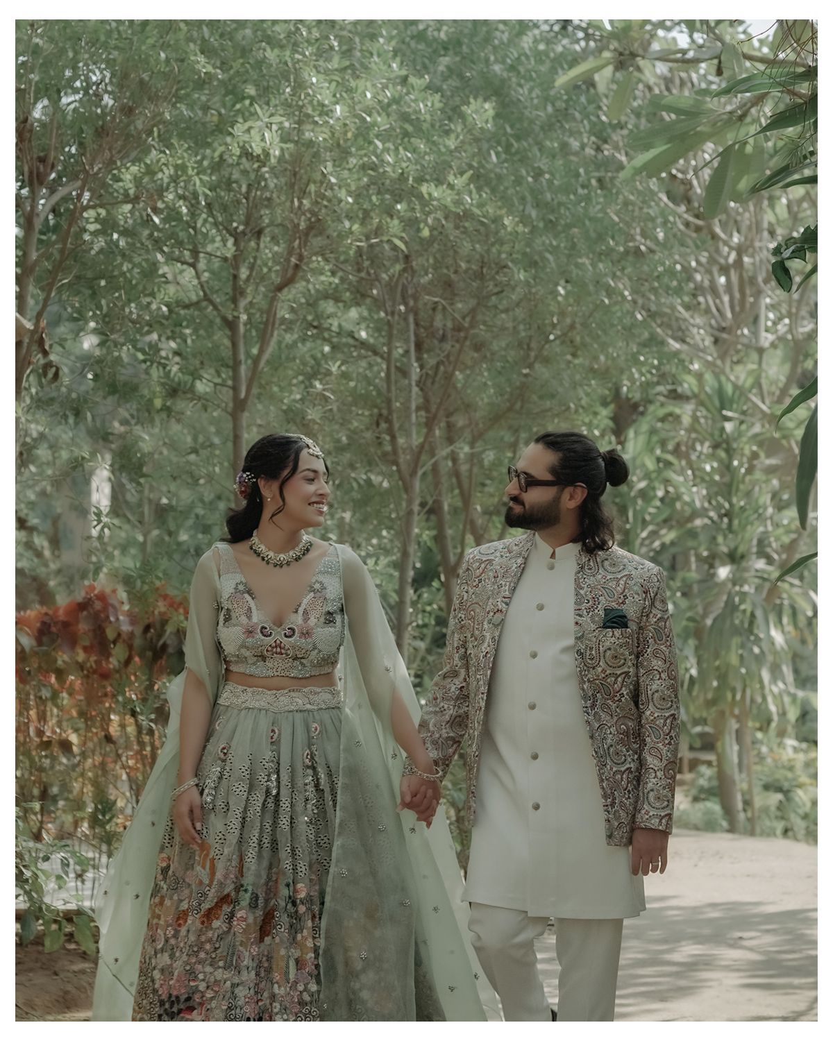 A couple in traditional Indian formal wear holding hands while walking on a tree-lined path in a garden setting.