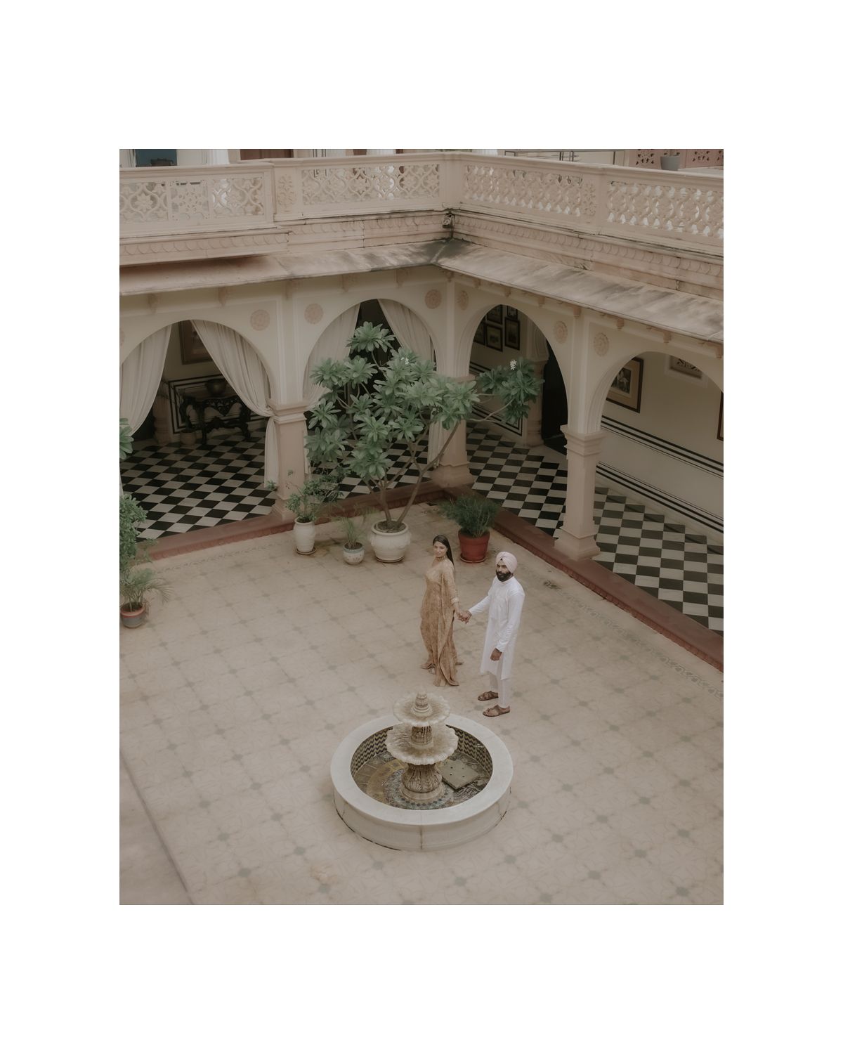 Aerial view of a colonial courtyard with checkerboard floor, central fountain, and two people conversing.