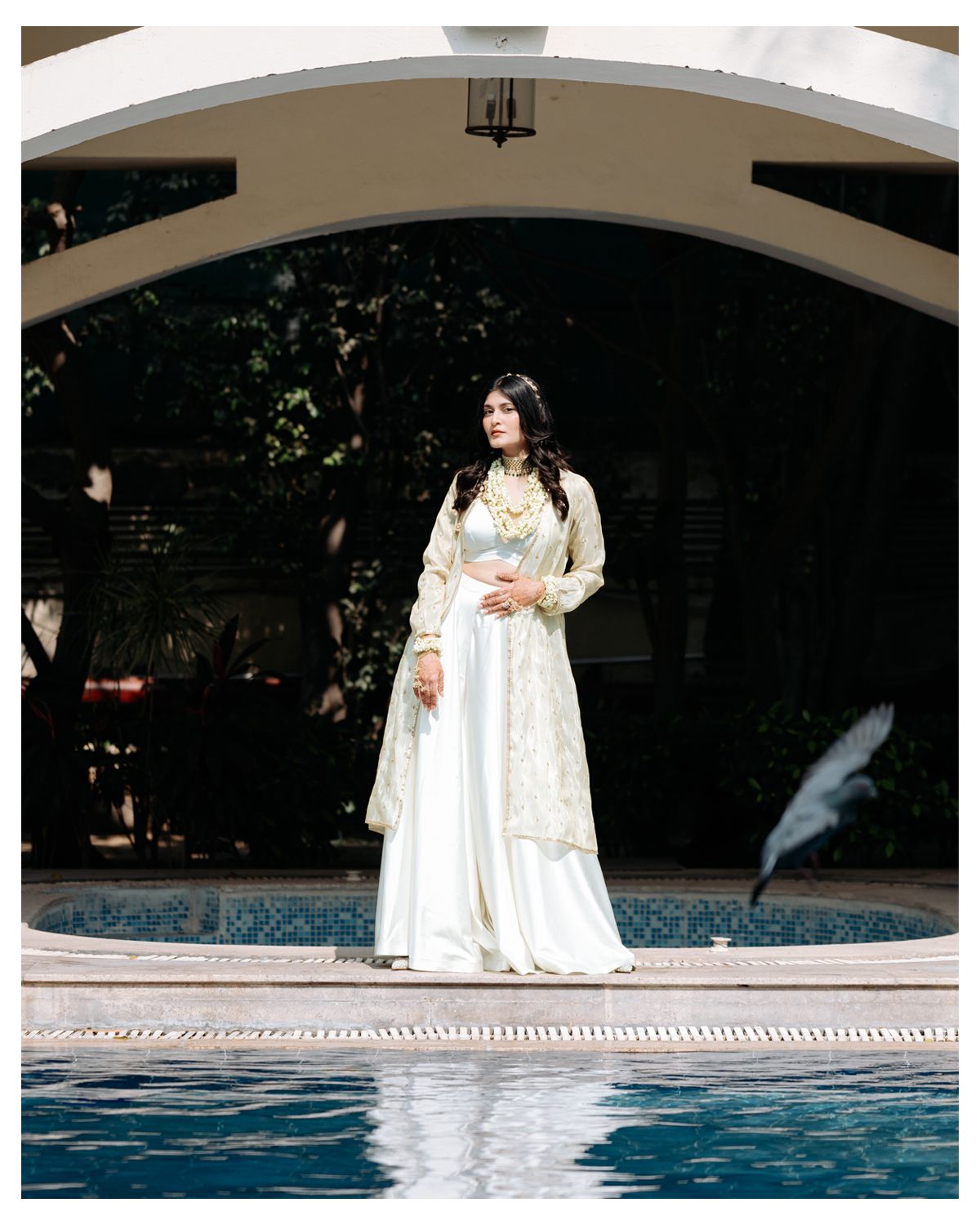 Bride in elegant white lehenga poses gracefully under an arched pavilion next to a swimming pool.