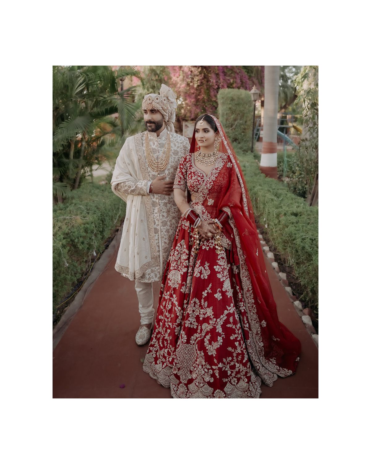 An elegant wedding couple poses on a garden path wearing traditional Indian bridal attire.