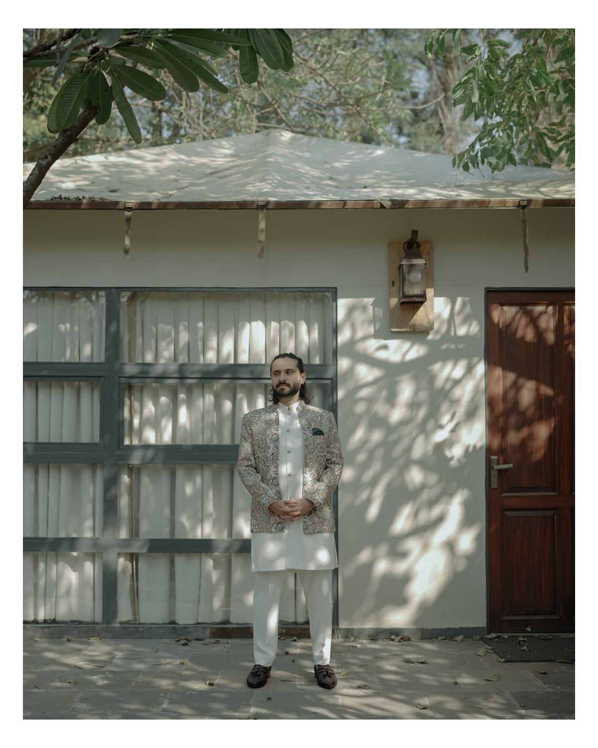 Someone in traditional Indian attire stands in front of a white house with dappled sunlight casting tree shadows on the wall.