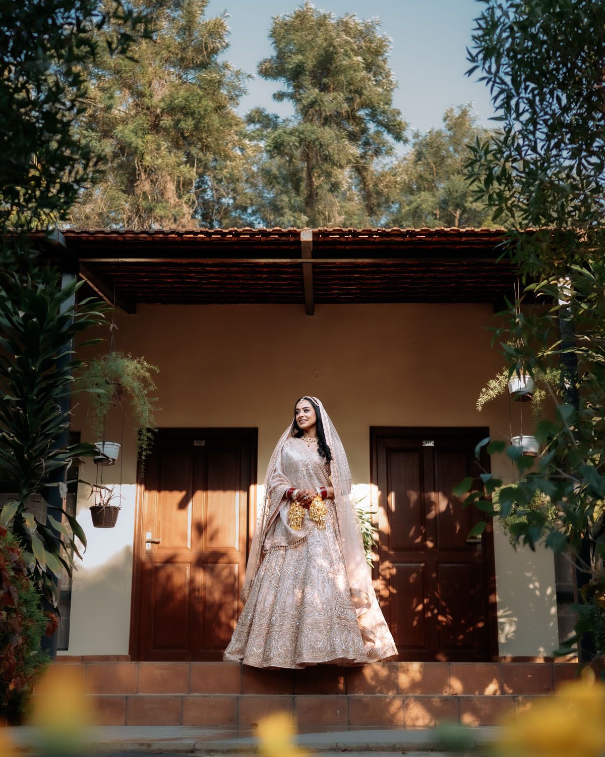 Person in an elegant white bridal gown stands on a covered porch with wooden doors and tall trees in the background.