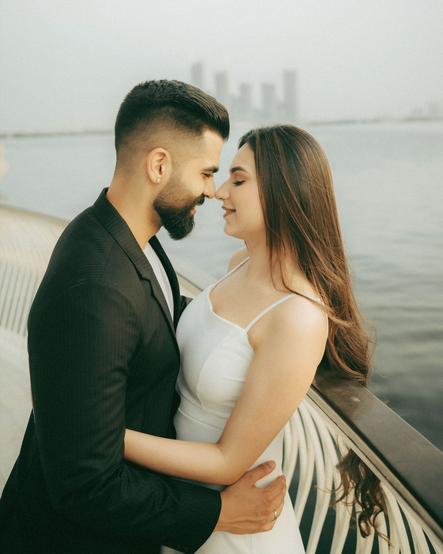 Romantic couple in formal wear sharing an intimate moment by the waterfront with city skyline in background.