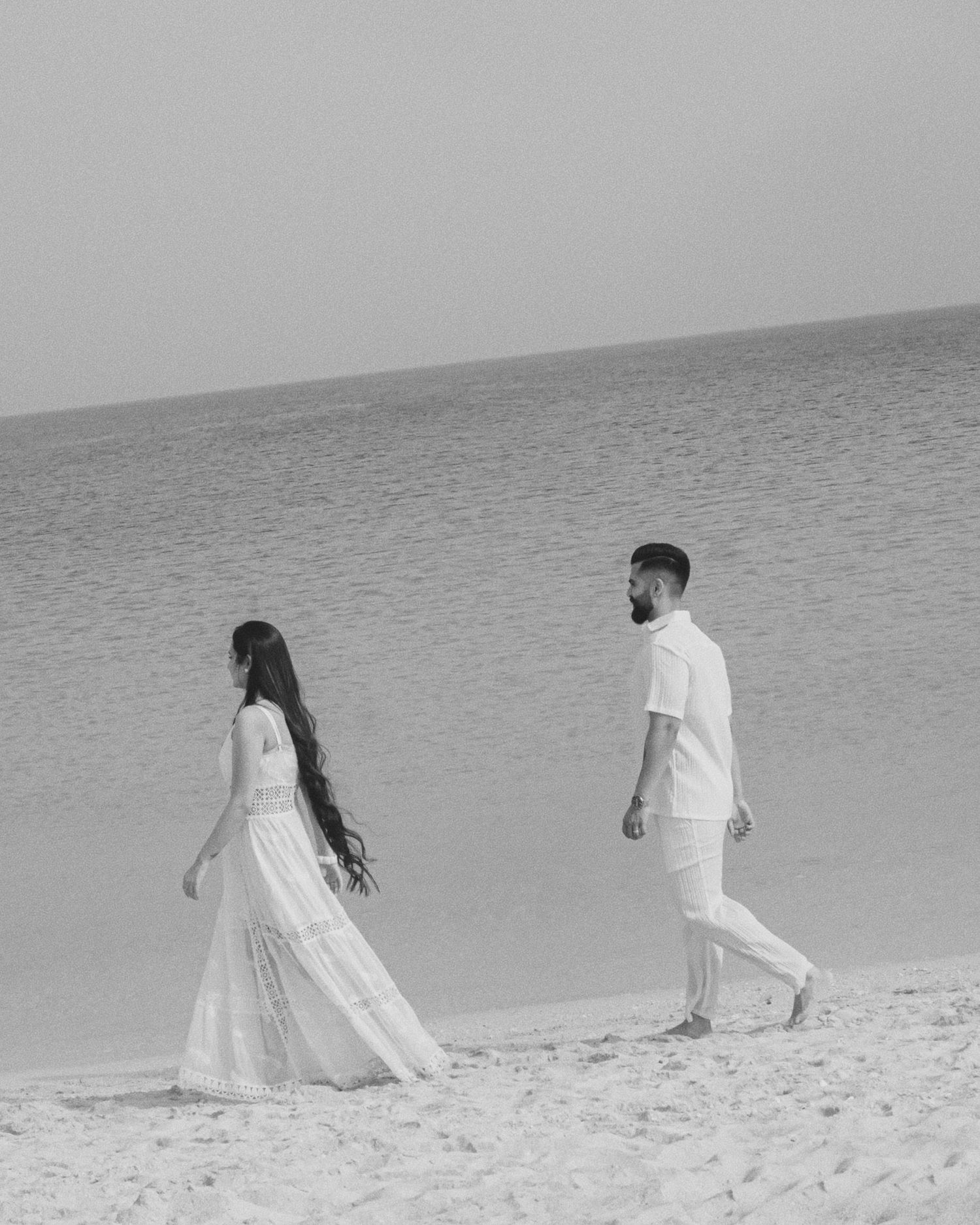 Black and white photo of a couple in white outfits walking on a sandy beach with calm ocean waters behind them.