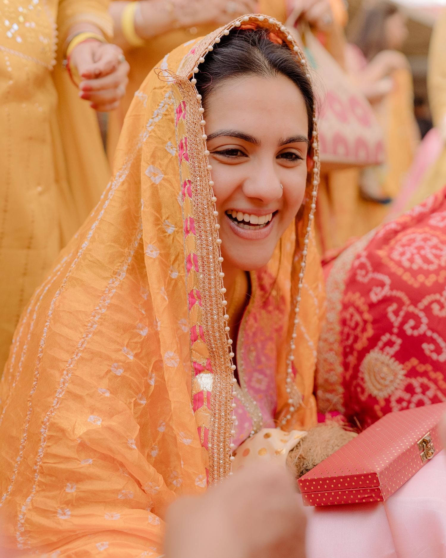 Smiling bride in orange embroidered dupatta and lehenga receiving gifts at a vibrant Indian wedding ceremony.