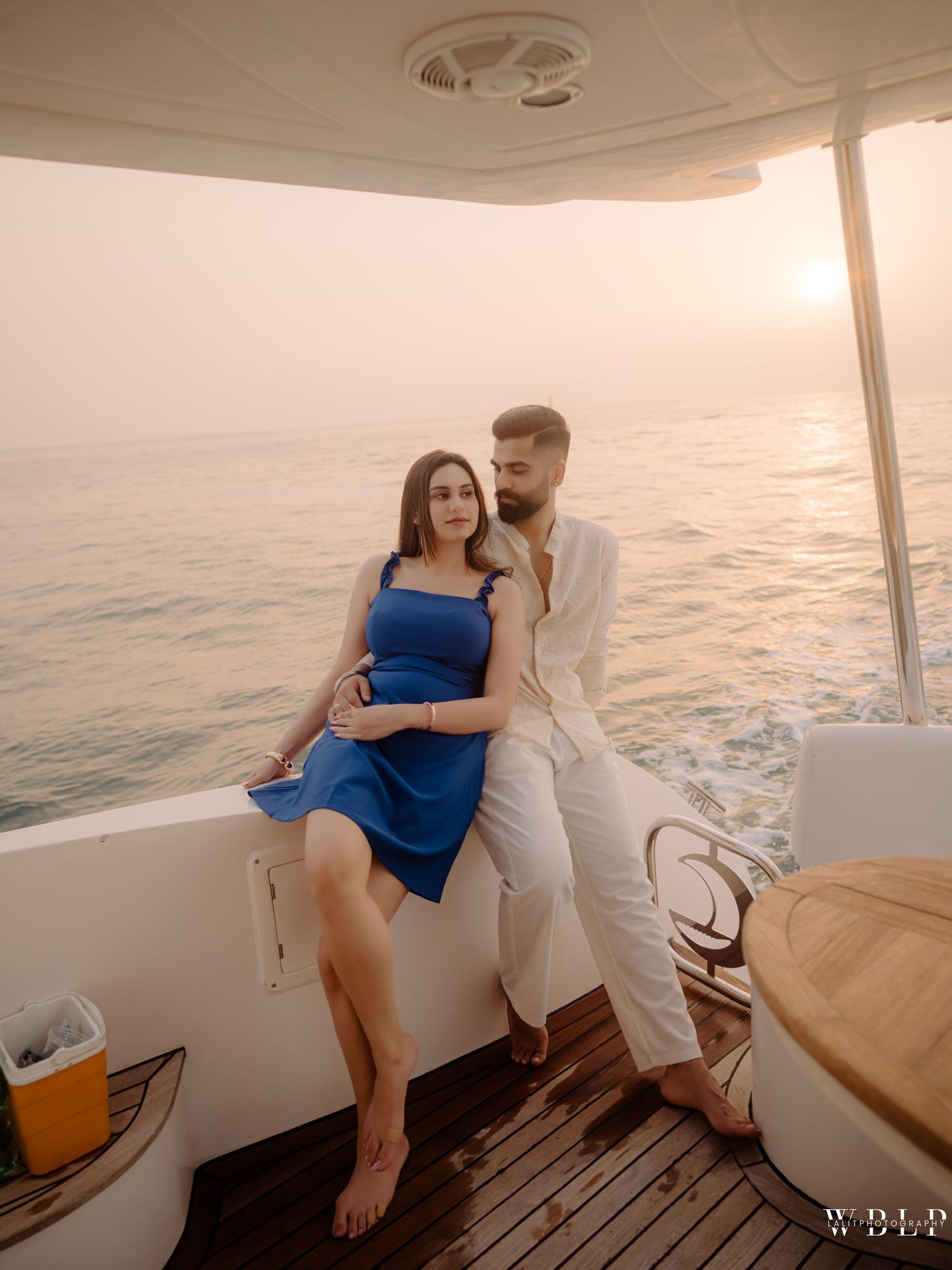 Couple posing romantically on a yacht at sunset, woman in blue dress seated, man in white outfit standing beside her.