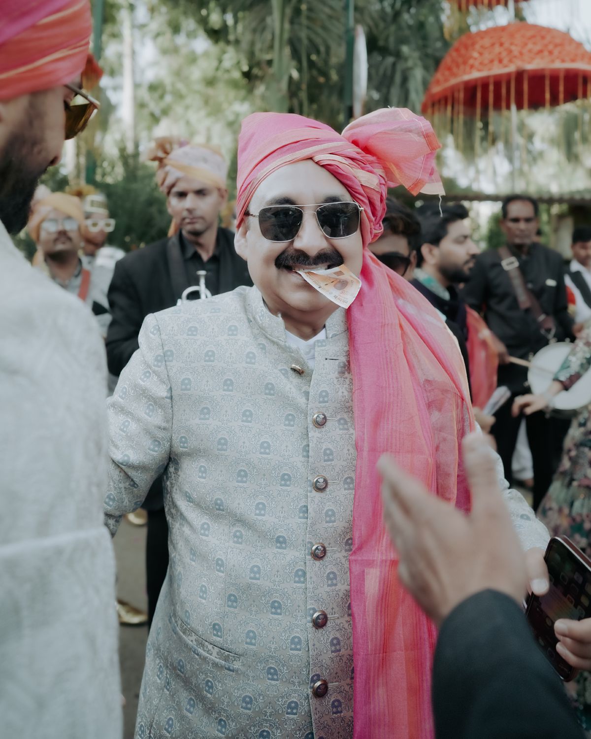 Wedding guest in traditional Indian attire with pink turban and sunglasses smiles at outdoor celebration.