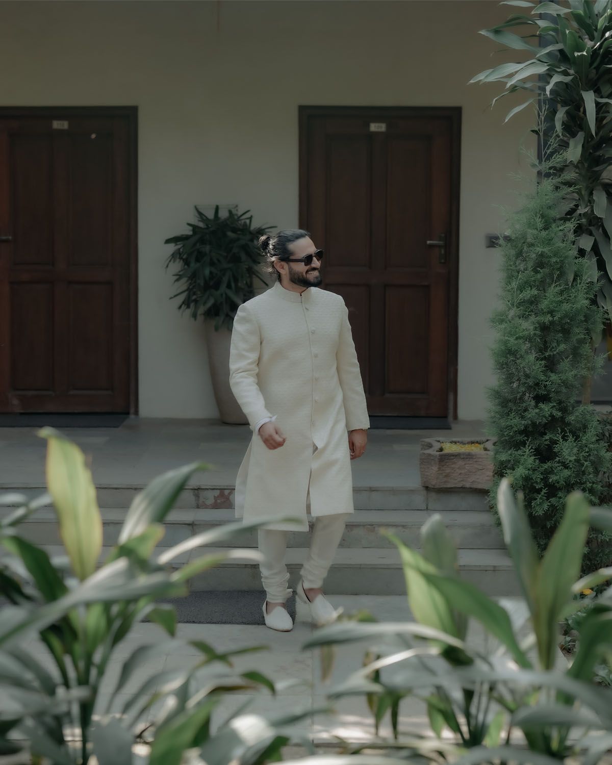 A person in a cream-colored traditional kurta and pants stands between wooden doors with tropical plants in the foreground.