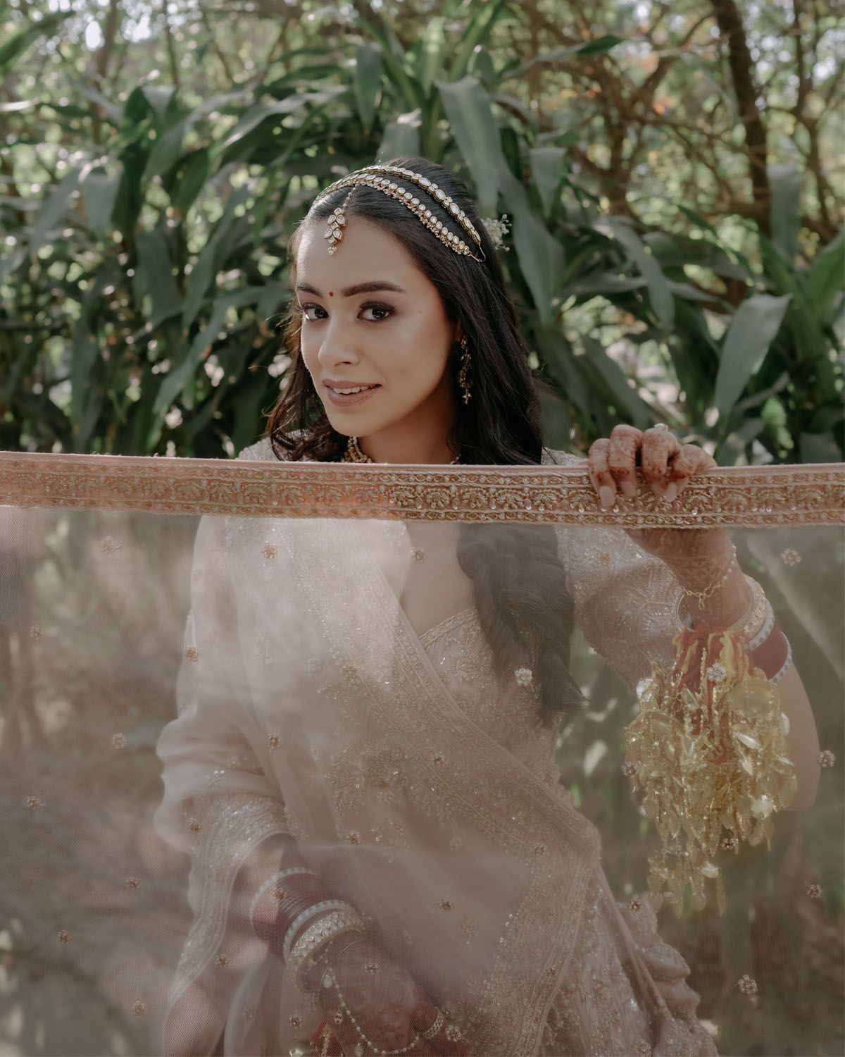 A beautiful South Asian bride in traditional attire poses behind sheer fabric in a garden setting with lush greenery.