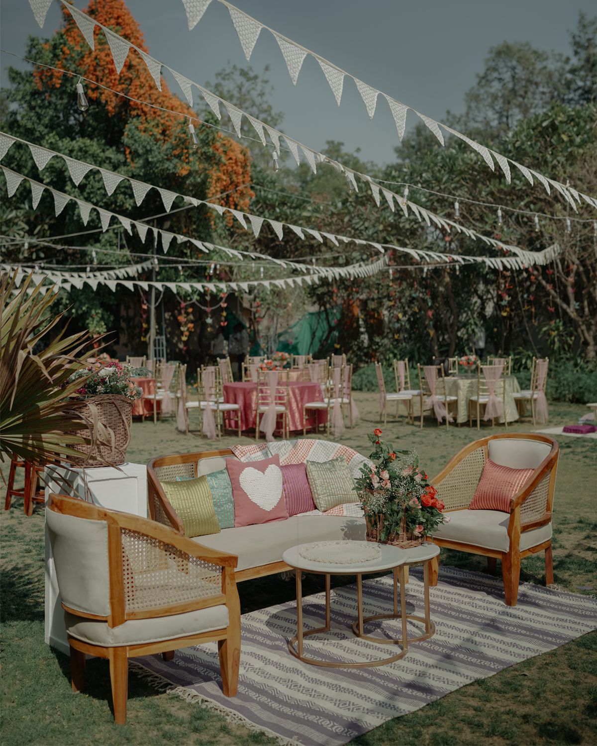 Outdoor wedding reception setup with white bunting decorations, rattan furniture, and round tables on a garden lawn.