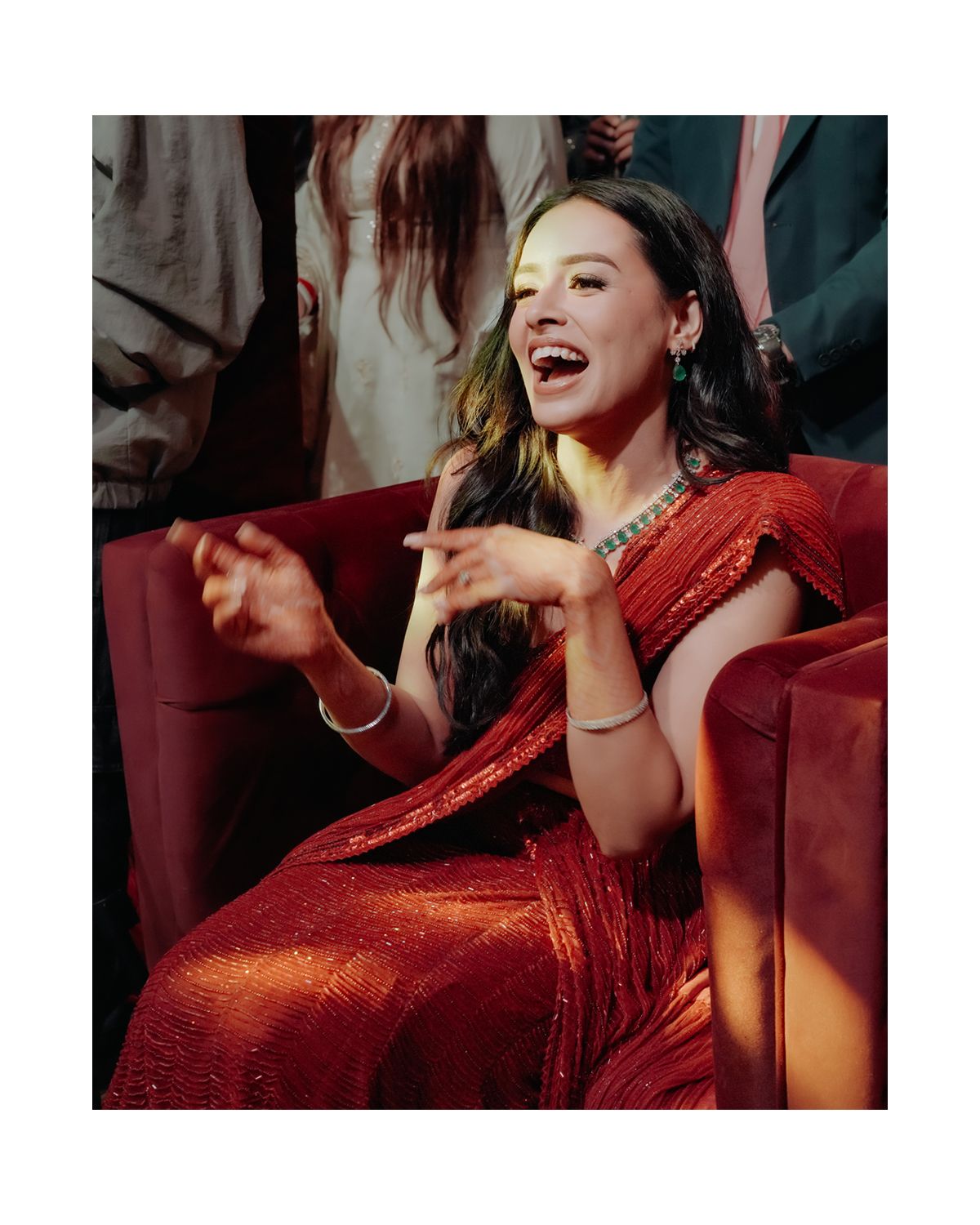 A laughing person in a red satin gown sits on a burgundy couch, gesturing expressively while being photographed.
