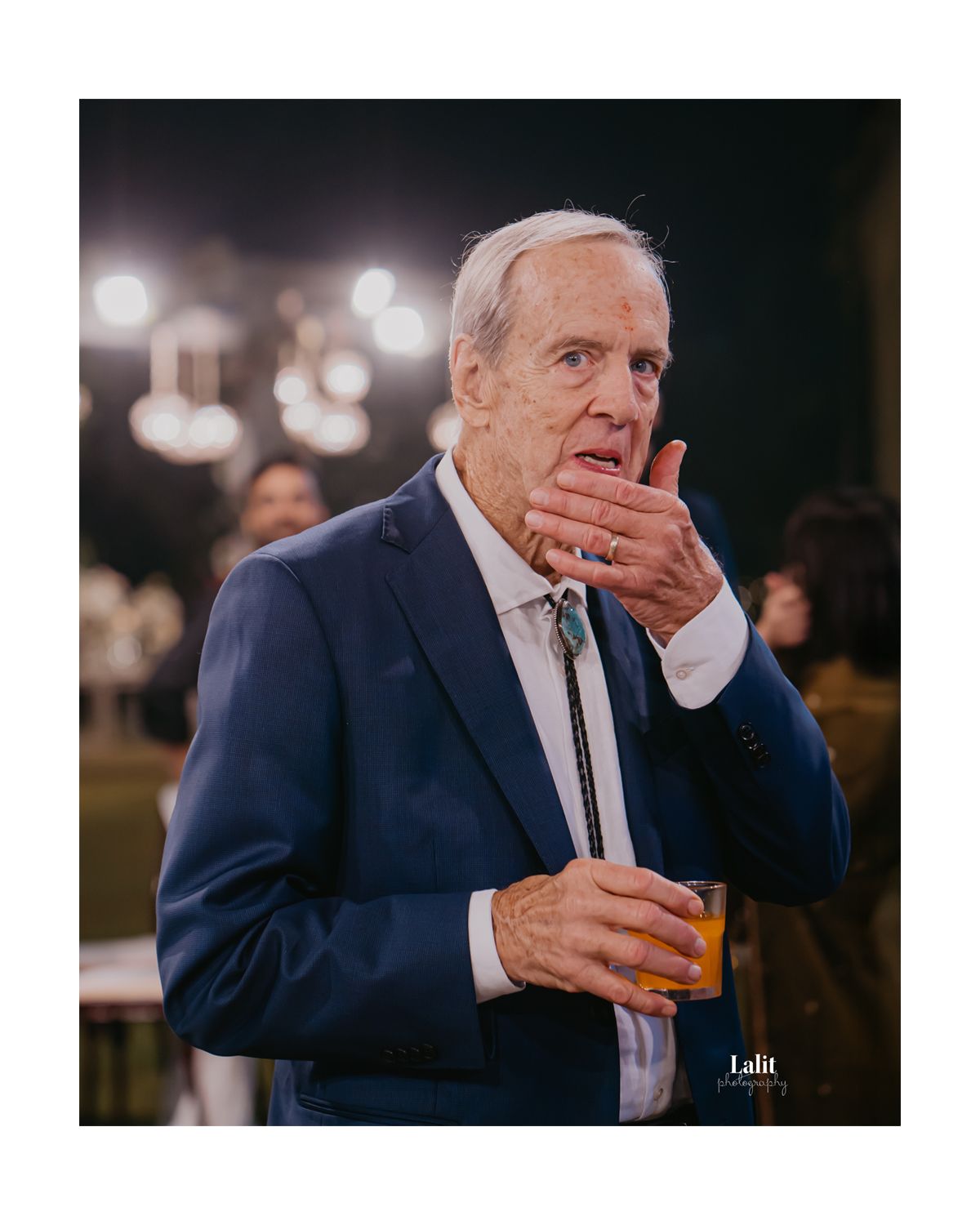 Groom's father in navy suit enjoys refreshments during an outdoor evening celebration.