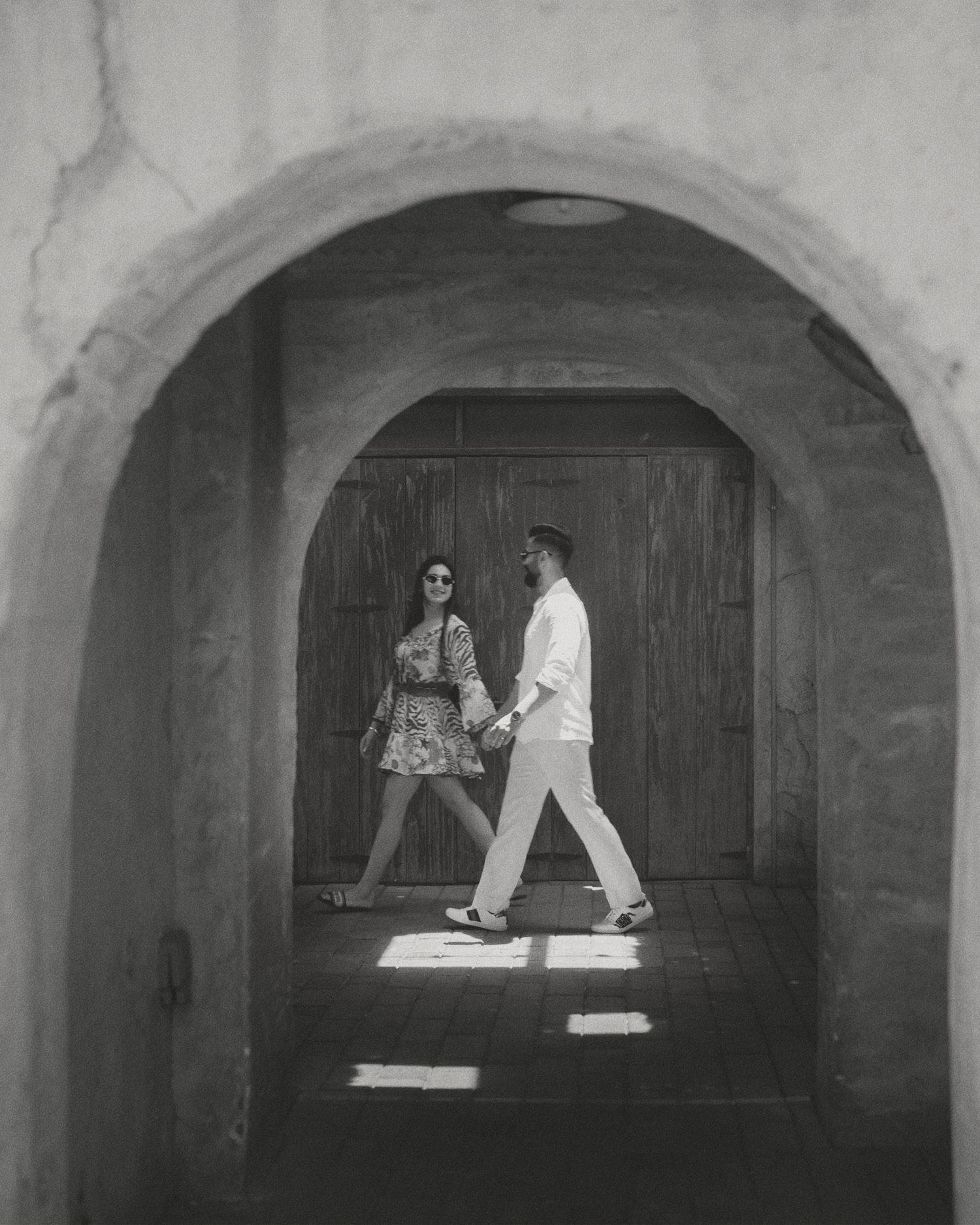 Black and white photo of a couple holding hands walking through an arched stone corridor in sunlight.