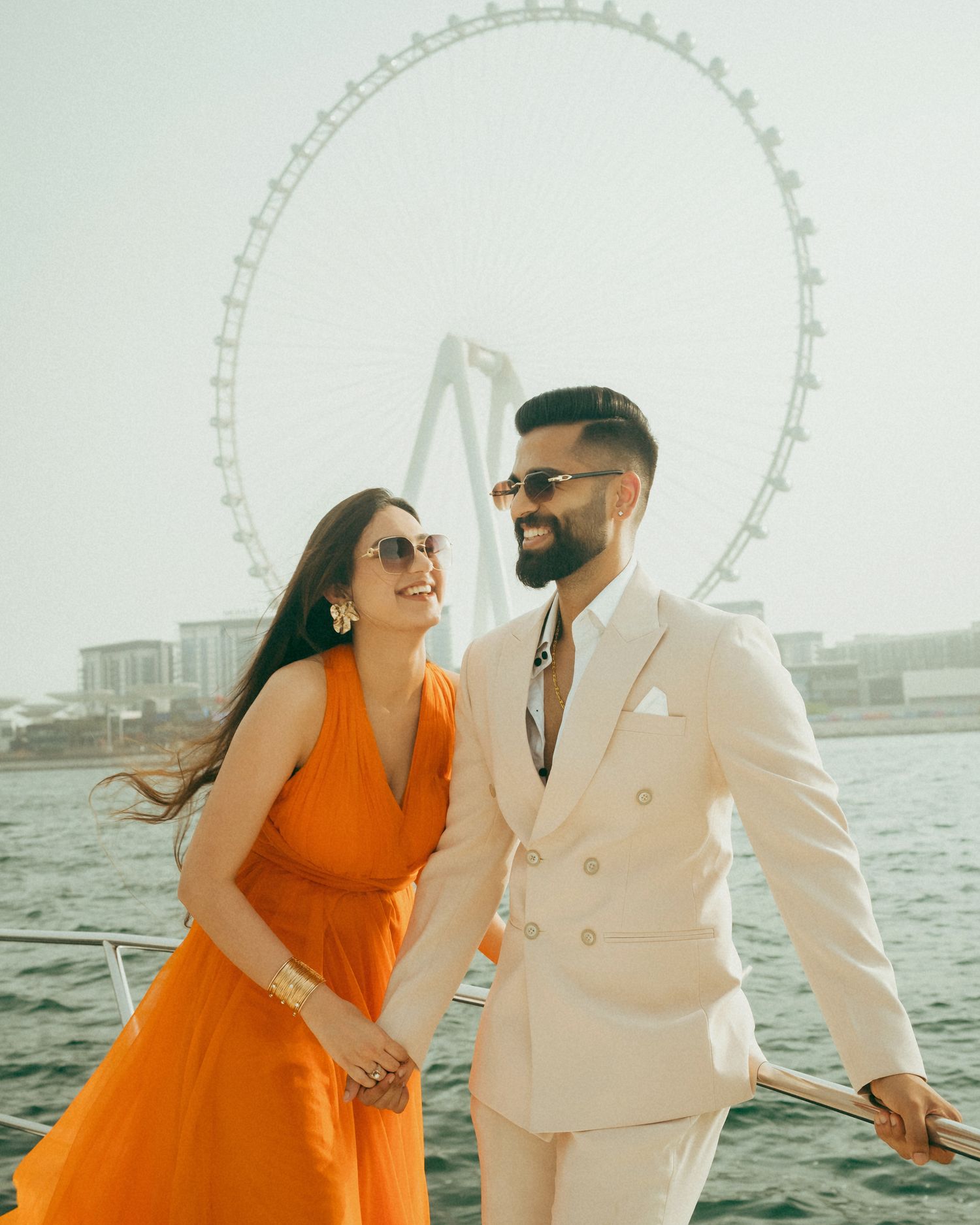 Couple smiling on a yacht with a large Ferris wheel in the background, woman in orange dress, man in white suit.