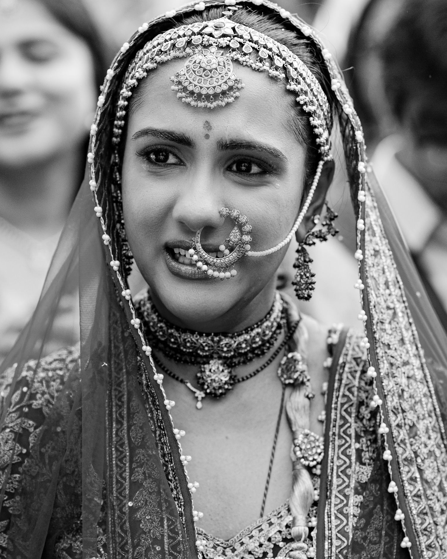 Black and white portrait of an Indian bride wearing traditional bridal jewelry, maang tikka, nose ring, and embroidered veil.
