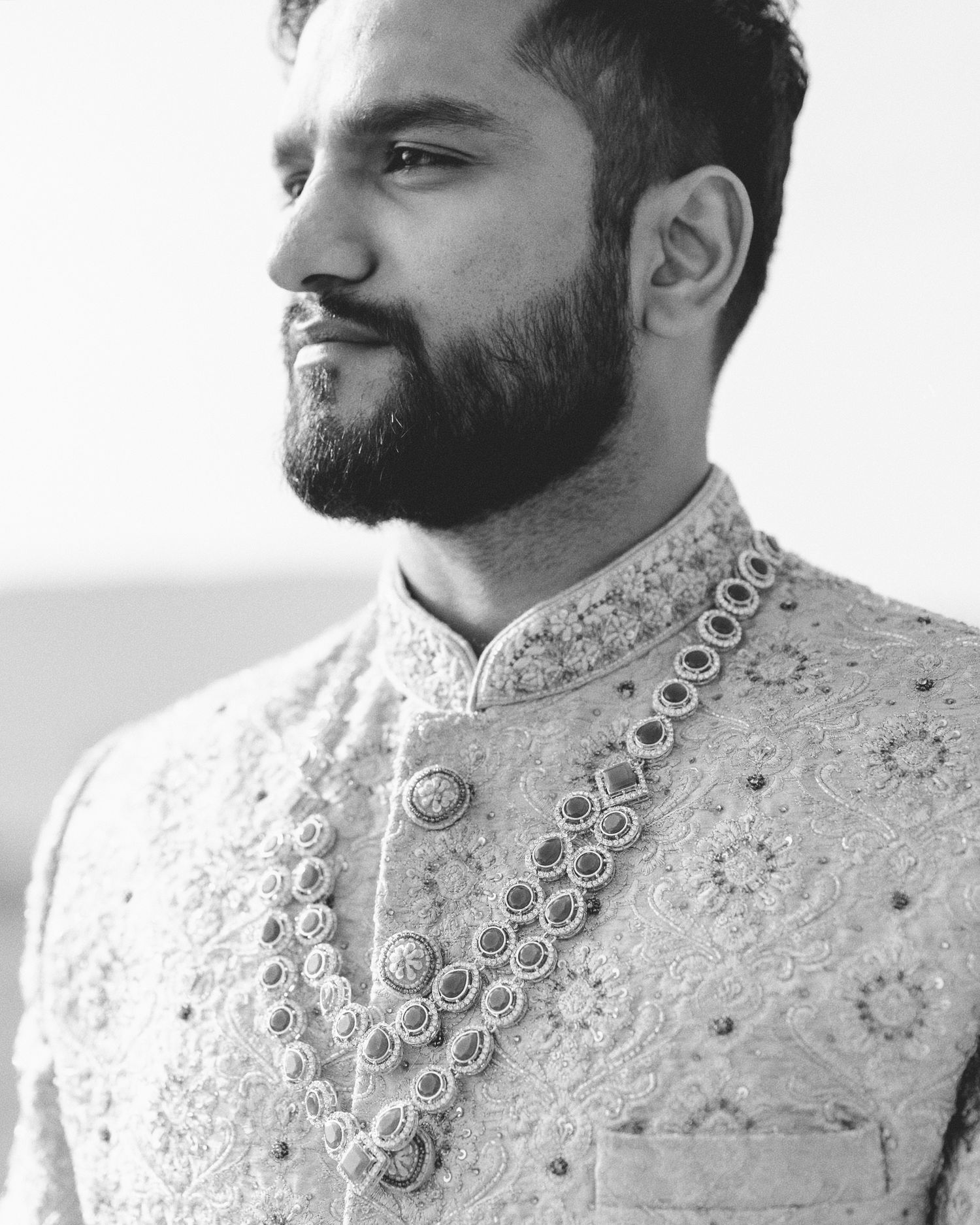 Black and white portrait of a bearded South Asian groom in embroidered sherwani with ornate layered necklace.