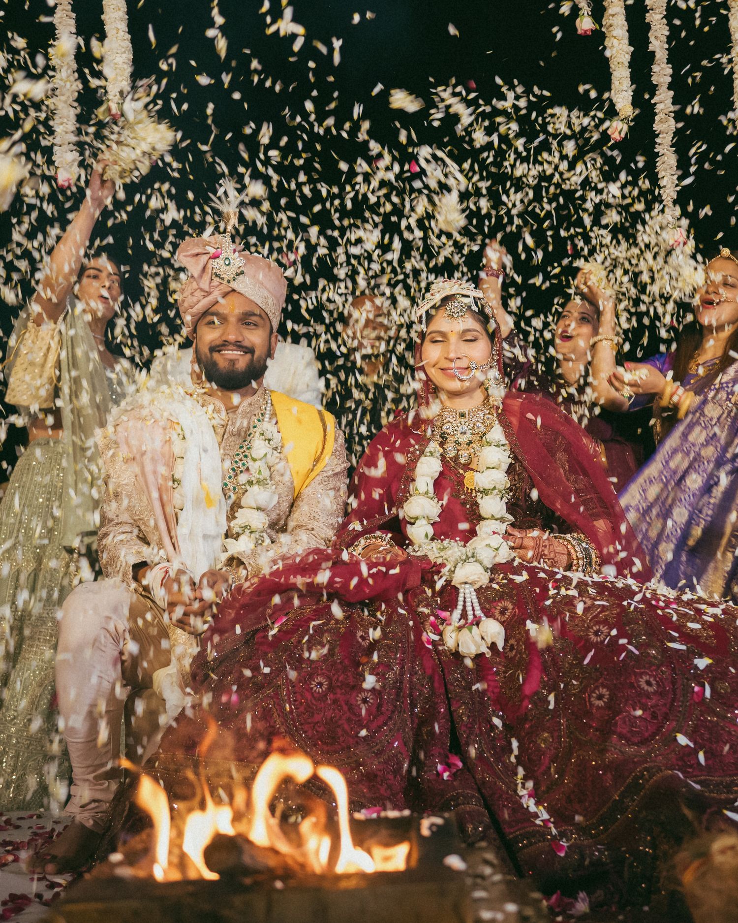 Indian bride and groom seated by sacred fire during traditional Hindu wedding ceremony with flower petals falling.