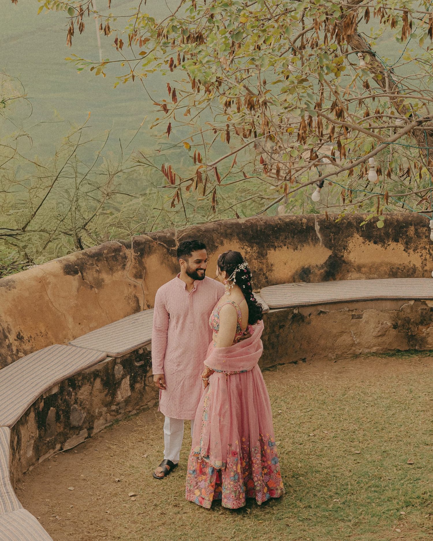 Indian couple in coordinating pink outfits sharing a romantic moment on a scenic terrace with trees and stone wall.