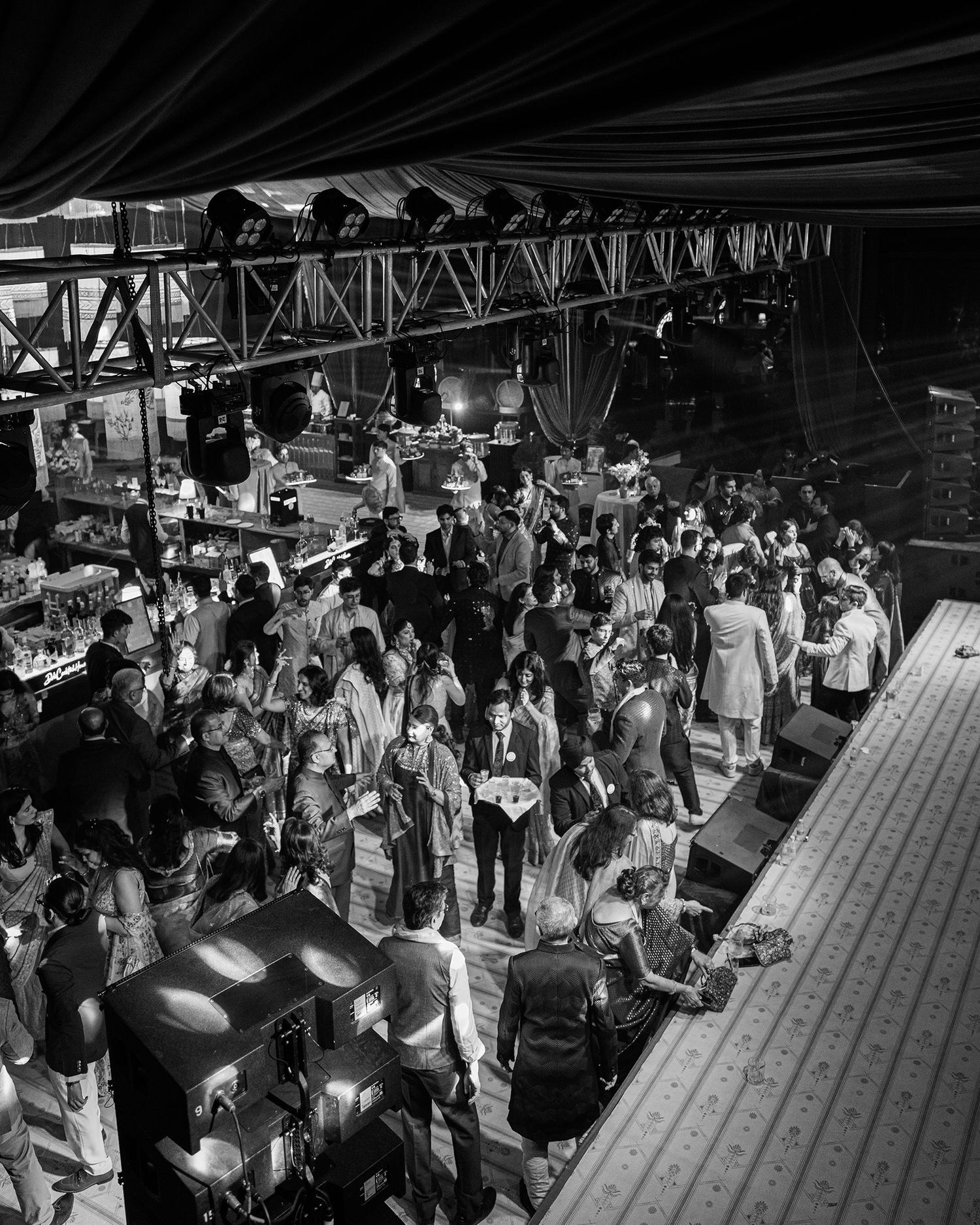 Black and white aerial view of a crowded indoor event venue with industrial steel beams and stage lighting overhead.