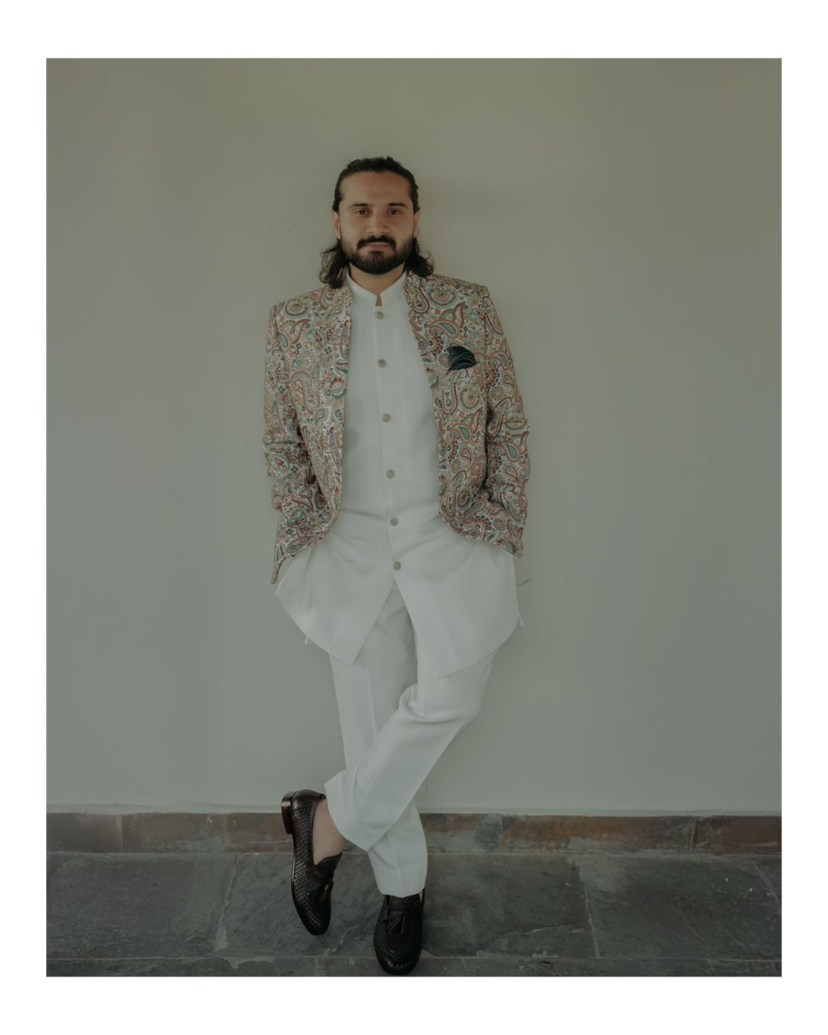 A person in a patterned beige blazer and white pants leans against a gray wall while wearing black dress shoes.