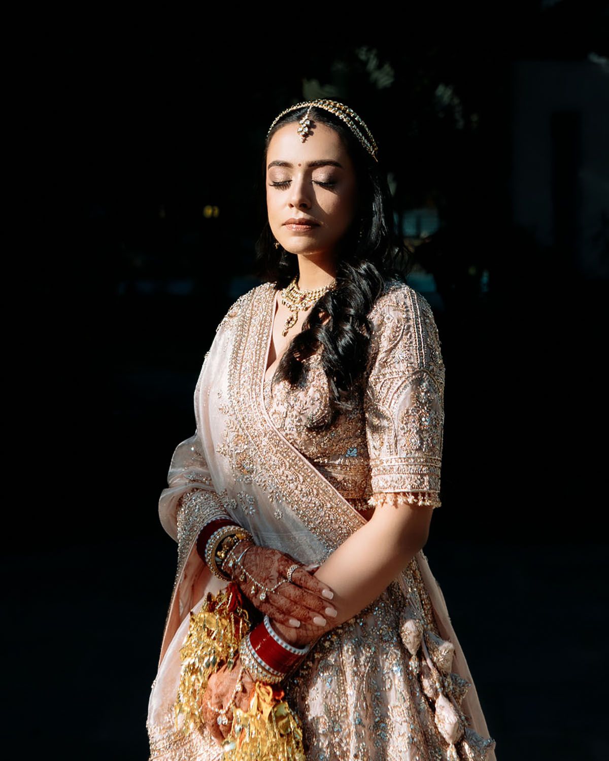 A bride in traditional South Asian attire with intricate beadwork and jewelry poses in dramatic lighting.
