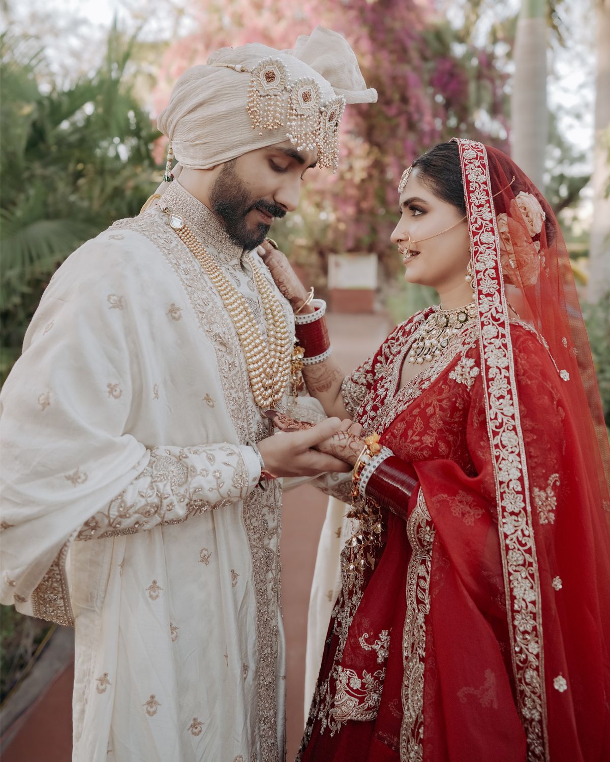 A romantic wedding portrait showing a couple in ornate Indian wedding outfits against a pink floral backdrop.