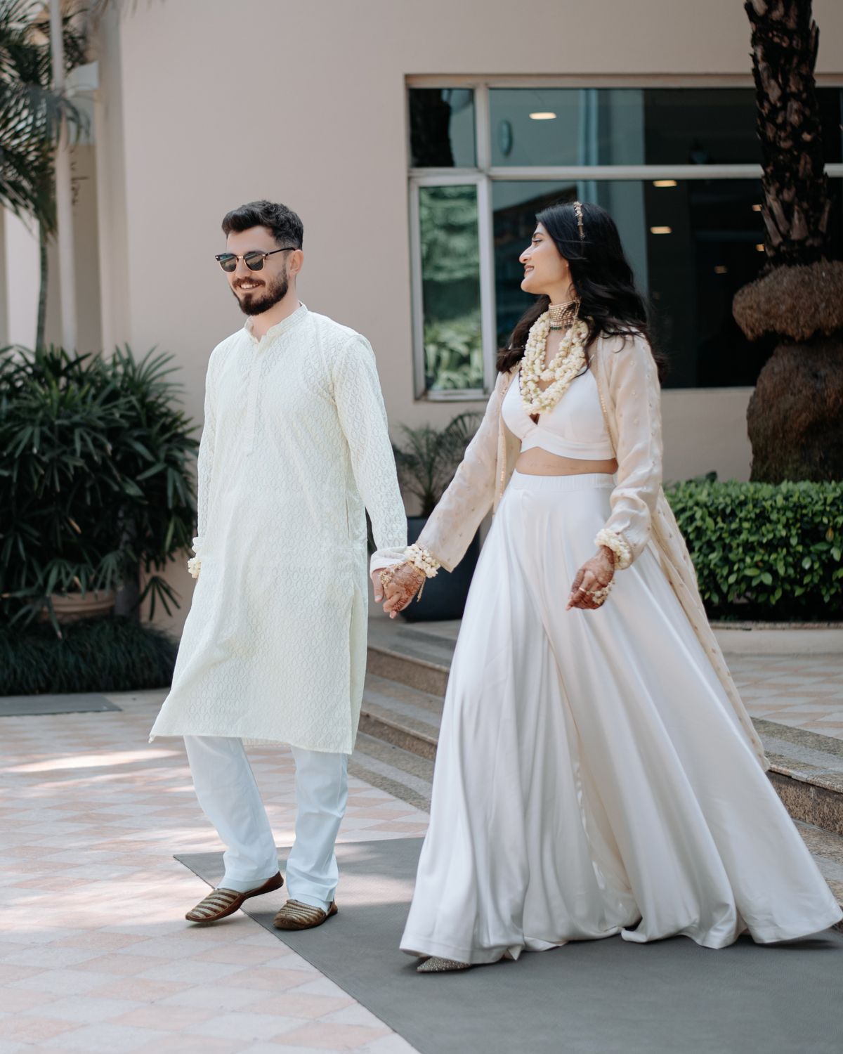 Couple walking hand in hand in elegant white traditional outfits through outdoor courtyard.