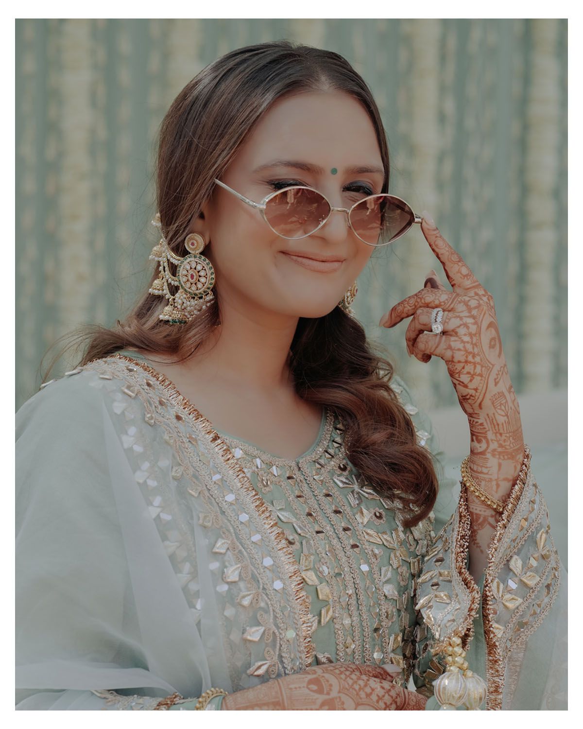 Person wearing traditional grey embroidered attire with henna-decorated hands and statement earrings.