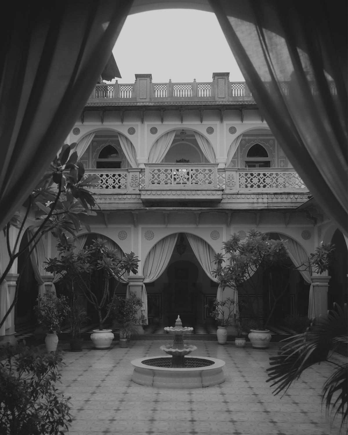 View through curtains of a colonial-style courtyard with fountain, potted plants, and ornate architectural details.