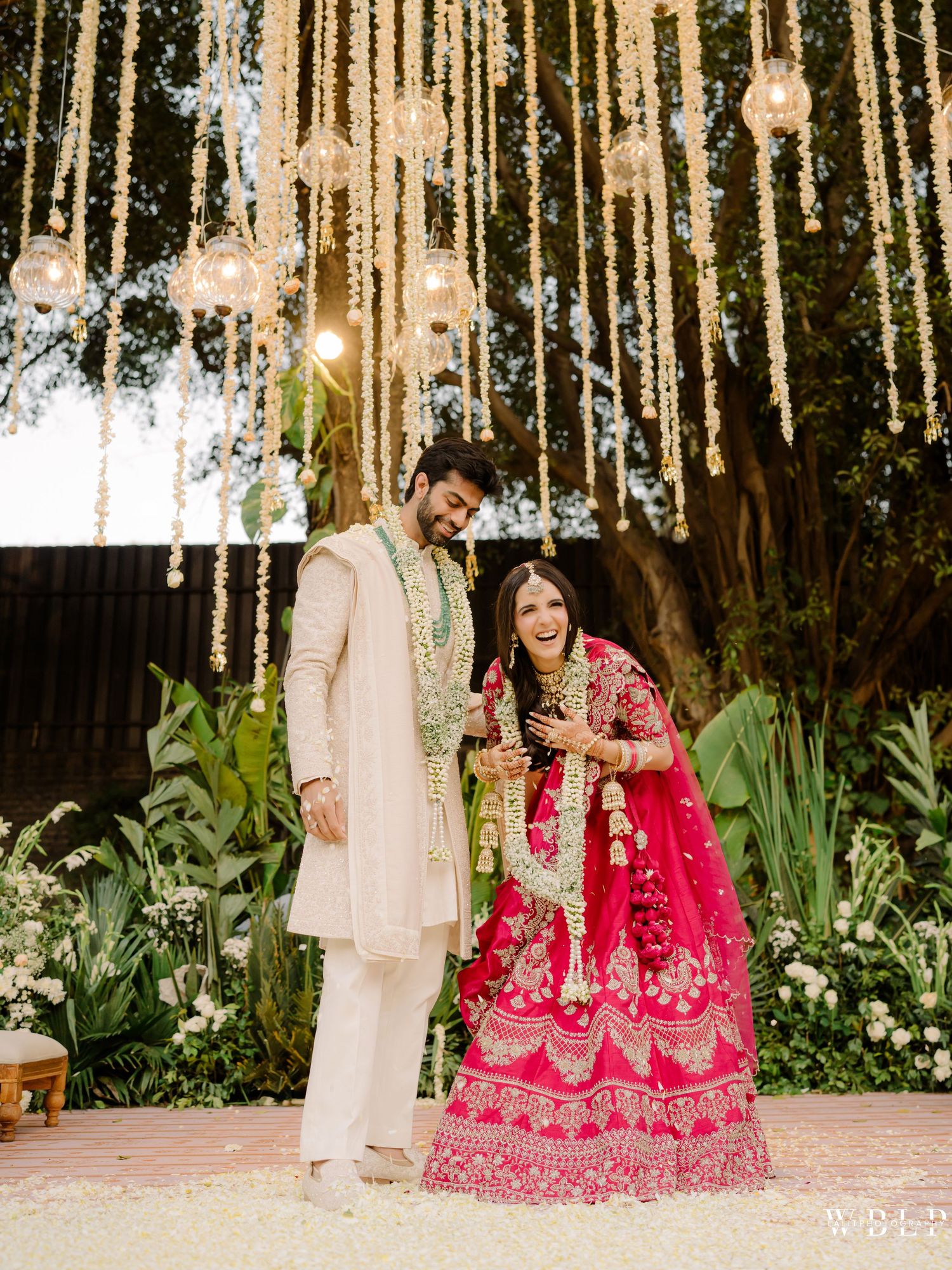 Couple in traditional Indian wedding attire smiling under floral chandelier backdrop with lush greenery.