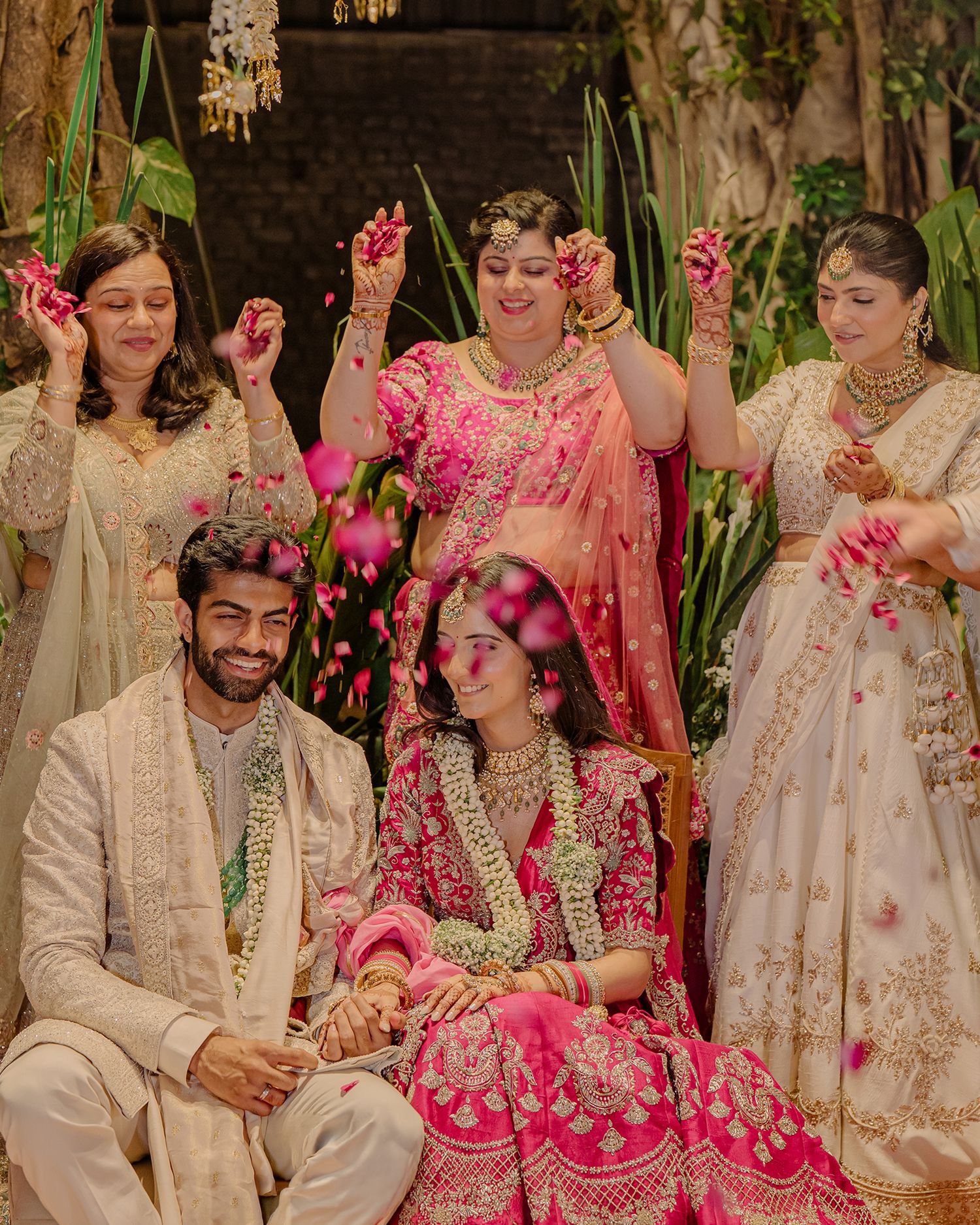Bride in pink lehenga and groom in white sherwani sit together as women shower flower petals at Indian wedding.