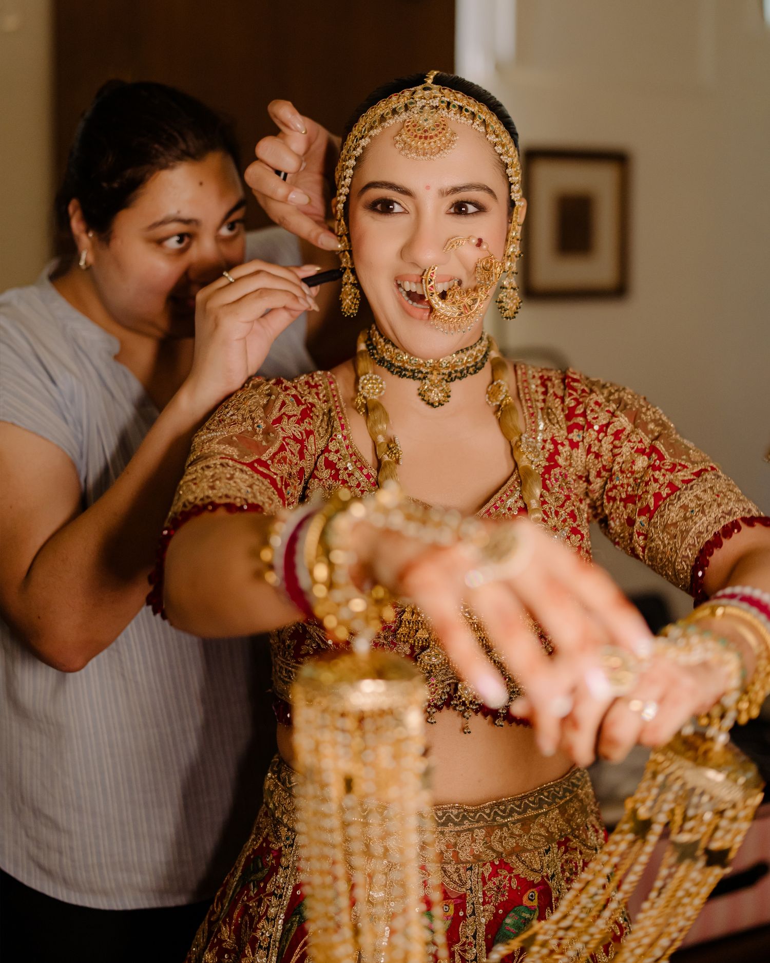 Indian bride in red and gold lehenga with traditional jewelry being assisted by makeup artist during wedding preparations.