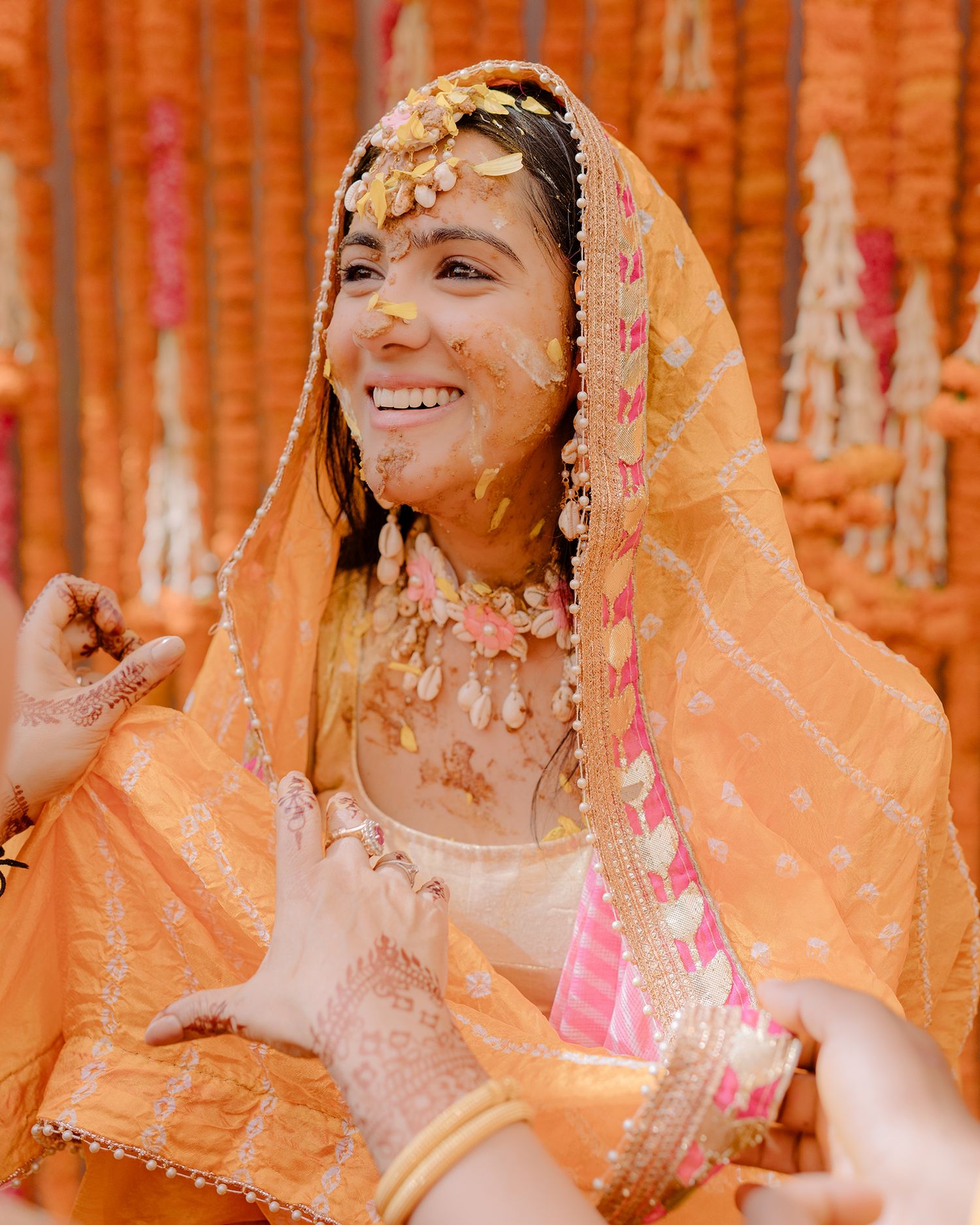Smiling bride in orange dupatta with haldi ceremony turmeric paste, wearing floral jewelry at Indian wedding.