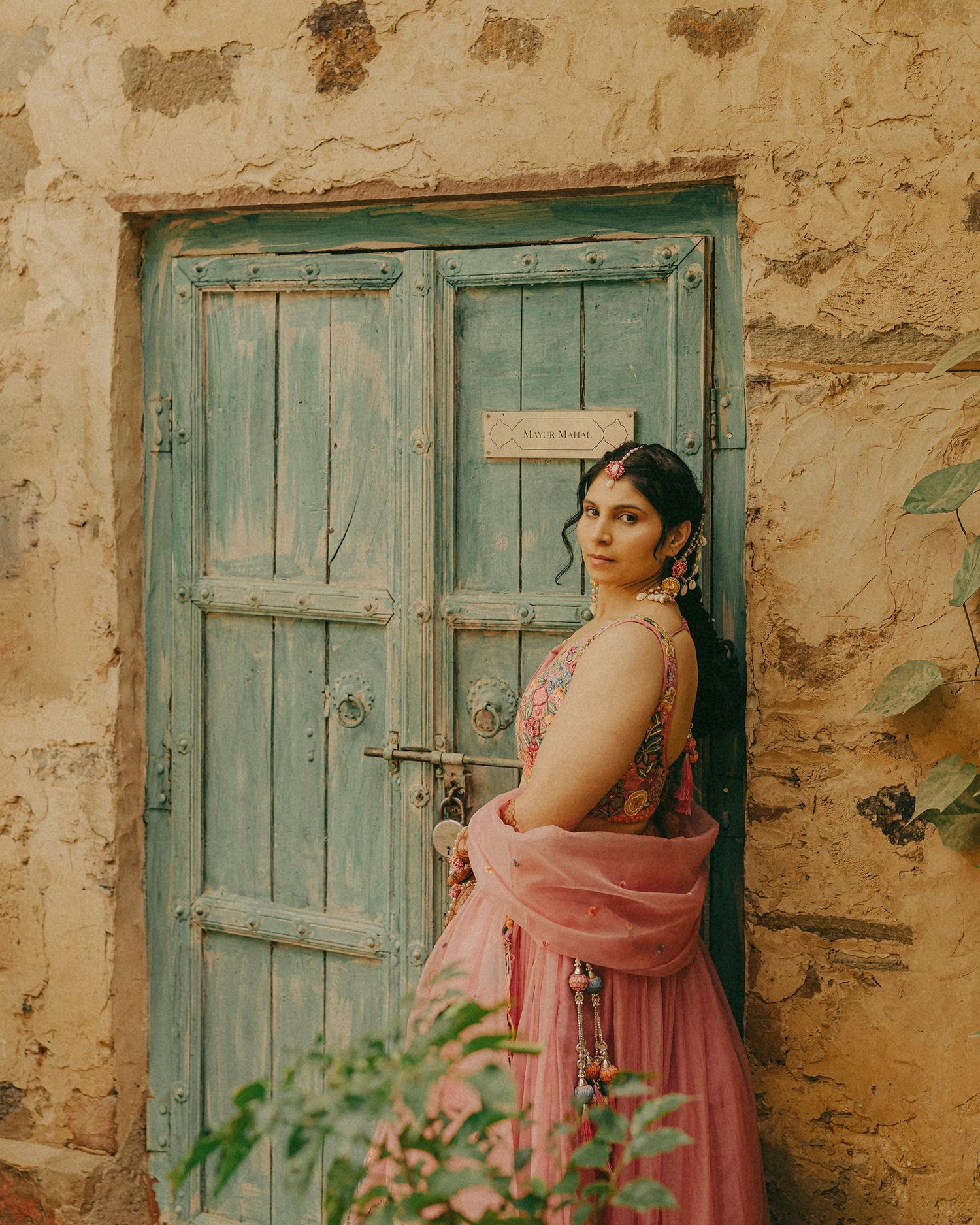 Woman in pink lehenga poses by rustic teal door in stone wall, wearing traditional Indian bridal attire.