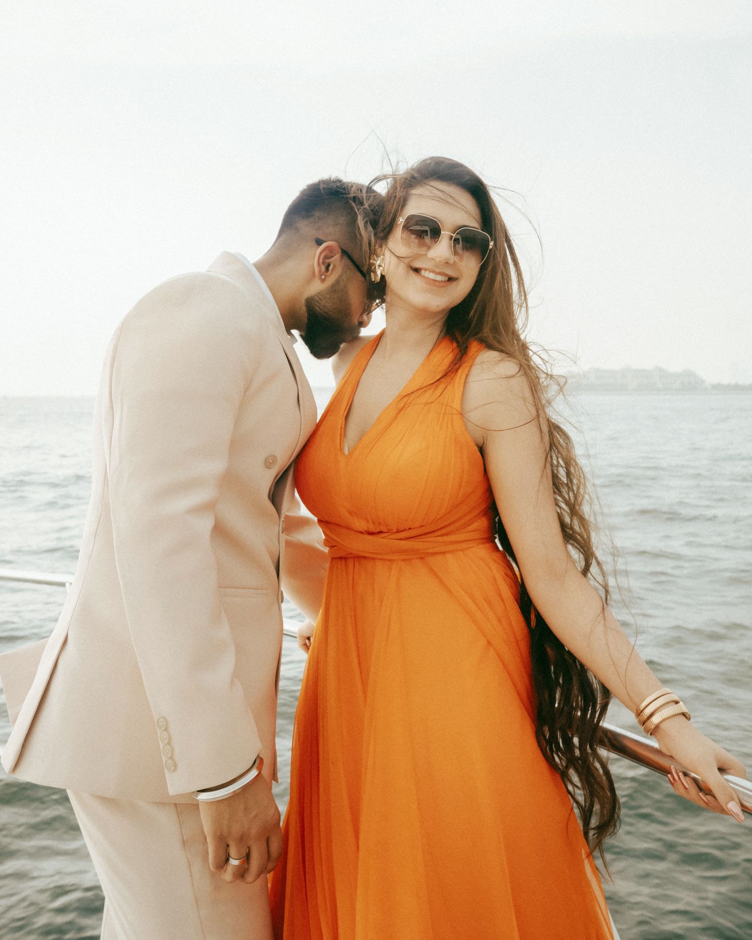 Couple posing on a boat, woman in orange dress with sunglasses, man in cream suit, ocean backdrop.