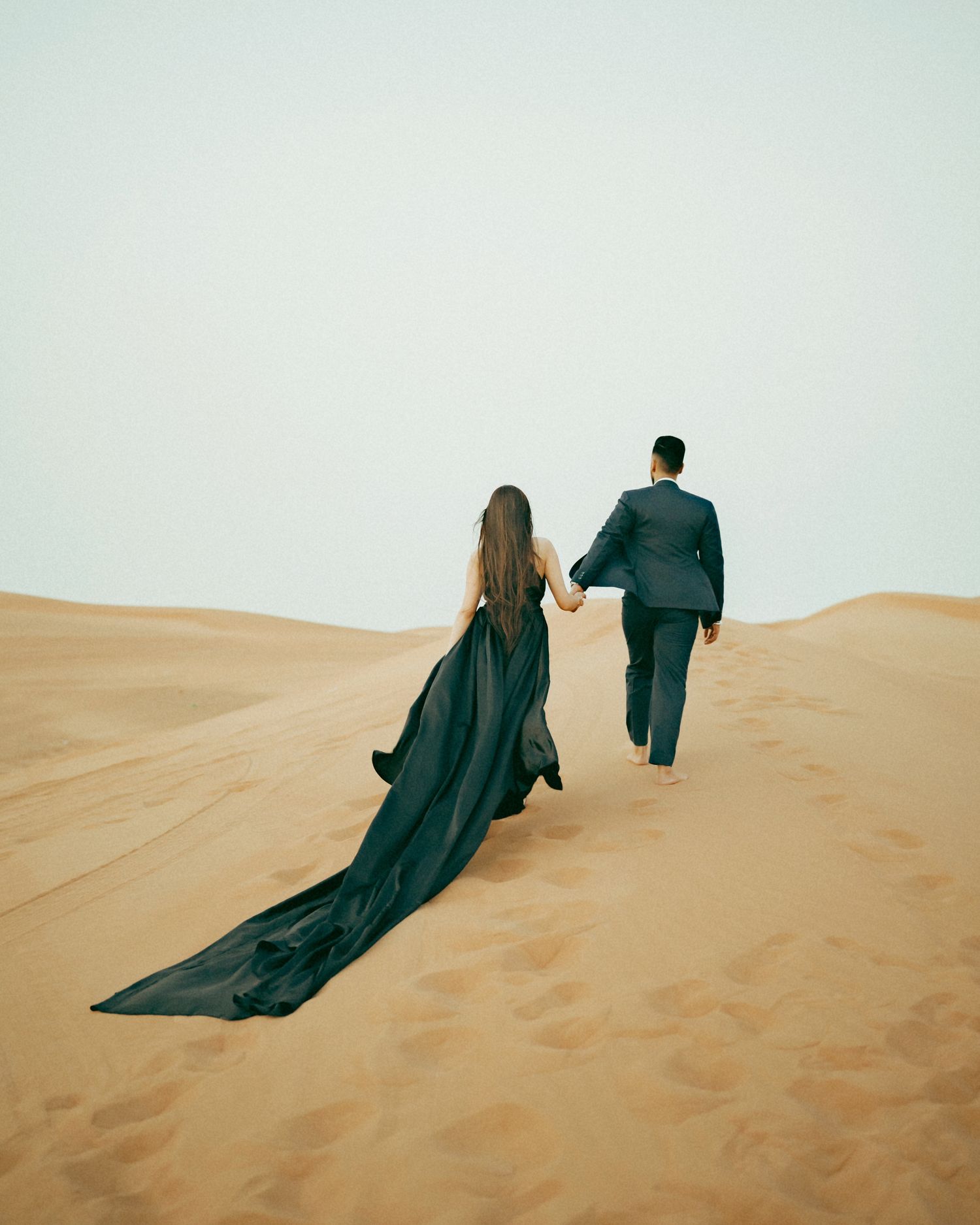 Couple in elegant dark attire walking hand in hand across golden sand dunes in a desert landscape.