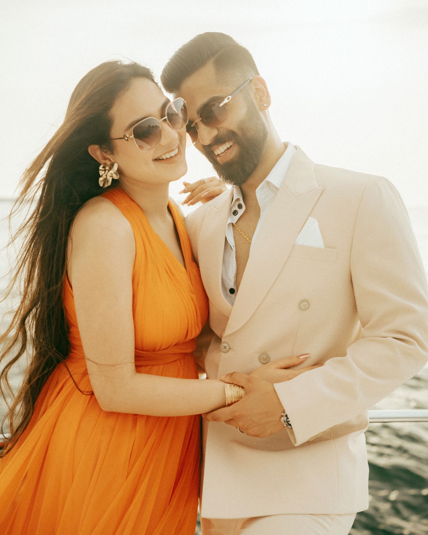 A couple in elegant attire, woman in orange dress and man in cream suit, smiling together outdoors in bright sunlight.