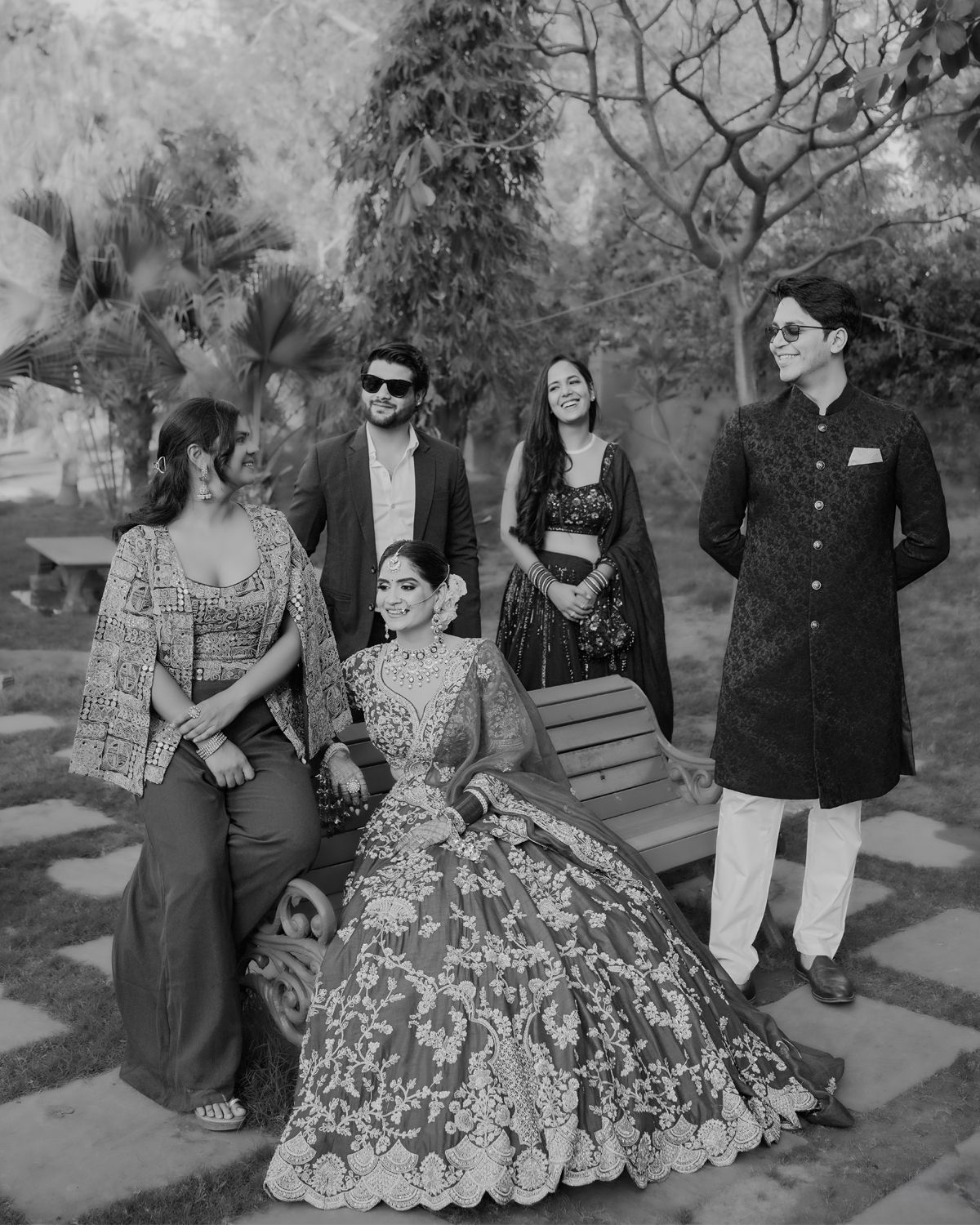 A group in traditional Indian attire poses outdoors in a garden setting with elegant formal wear and lehengas.