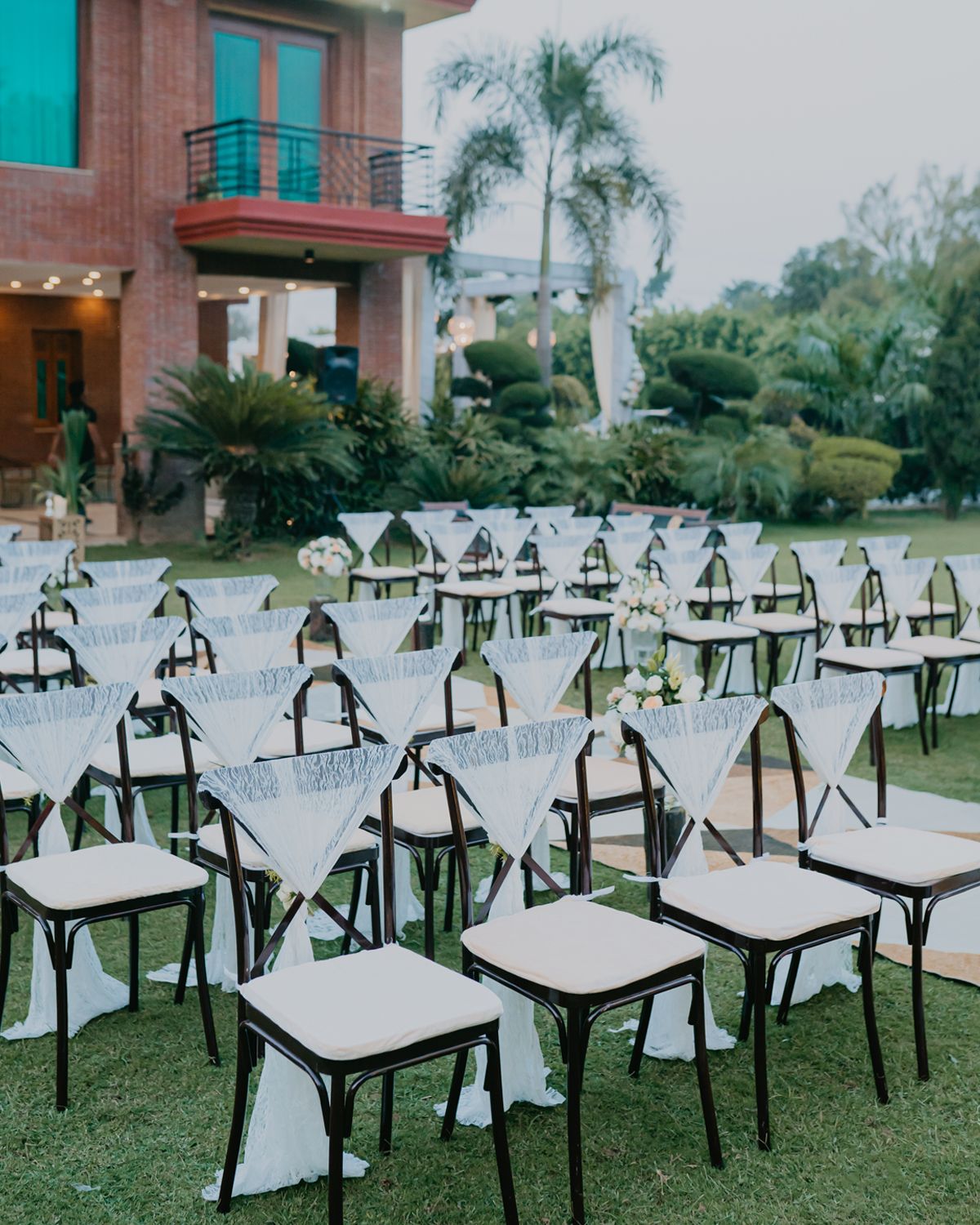 White chairs arranged in rows on a lawn with palm trees and a brick building in the background.
