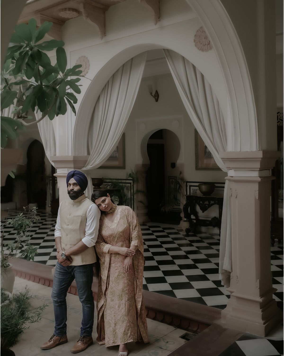 Two people posing beneath grand white archways with checkered flooring and plants.
