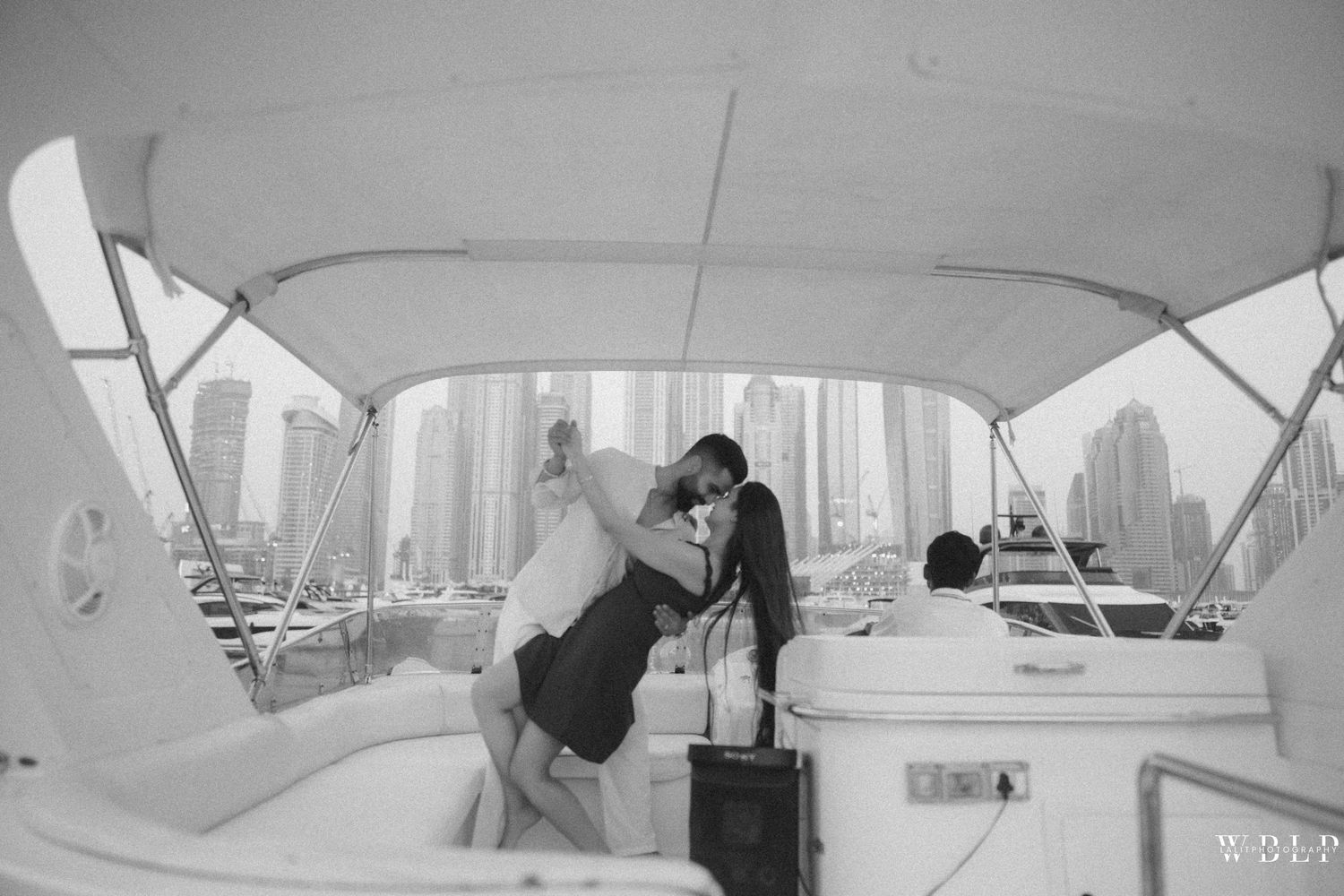 Couple embracing on a boat deck with city skyline in background, black and white wedding or engagement photo.