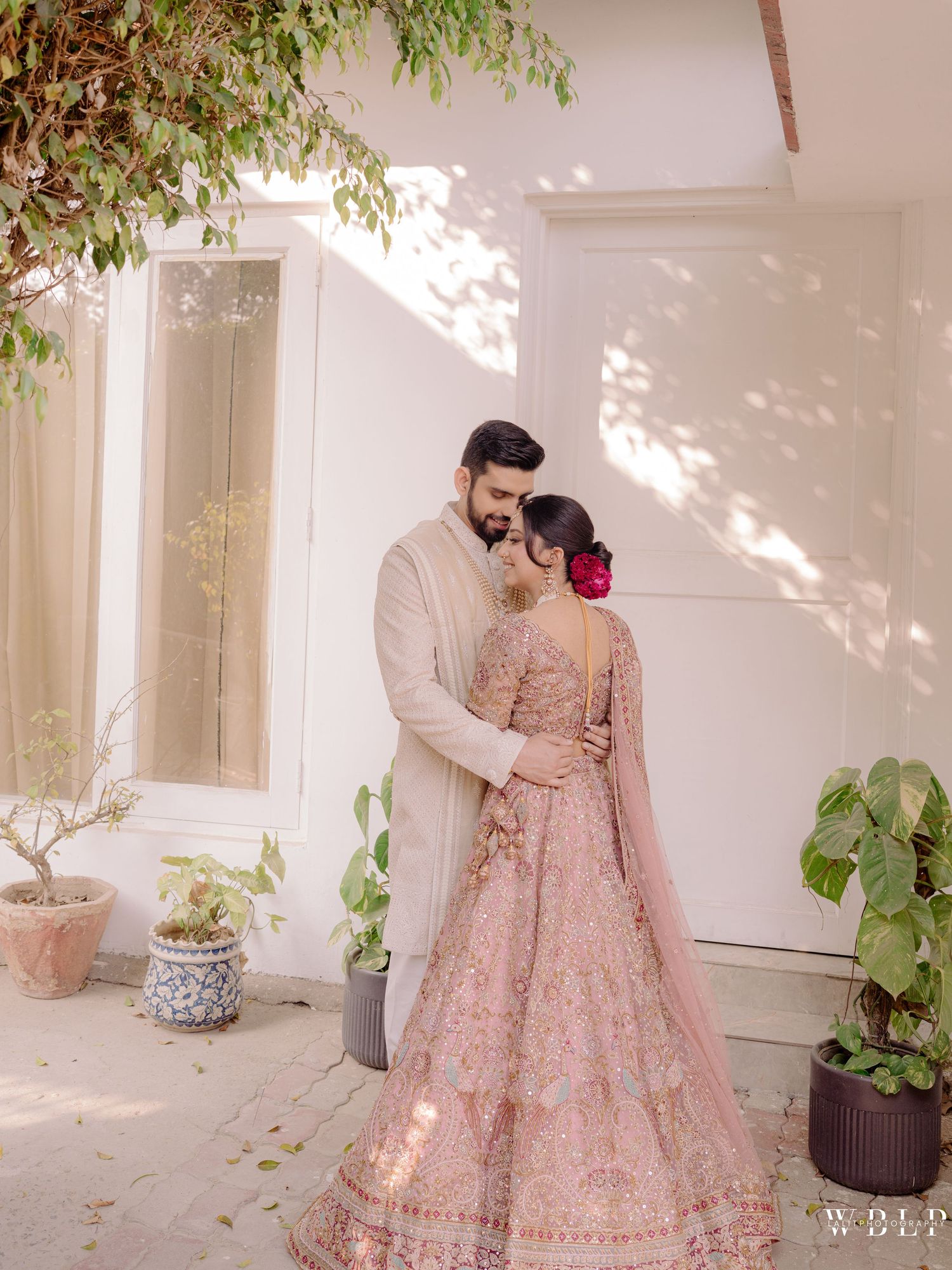 Couple in elegant Indian wedding attire, bride in pink lehenga with floral hair, groom in beige sherwani, embracing outdoors.
