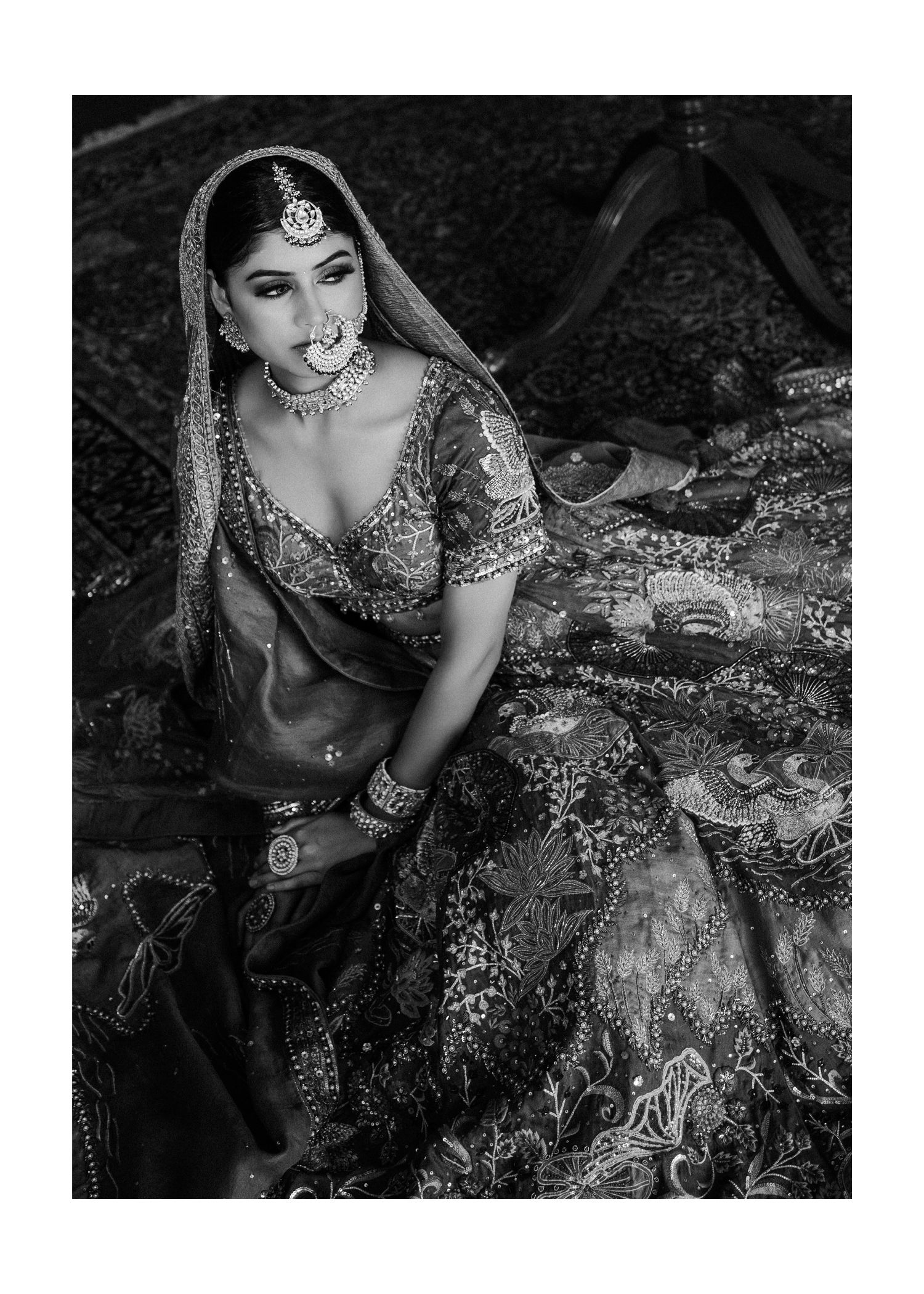 Bride in traditional Indian attire featuring intricate embroidery and jewels poses against a dark background in a monochrome photo.