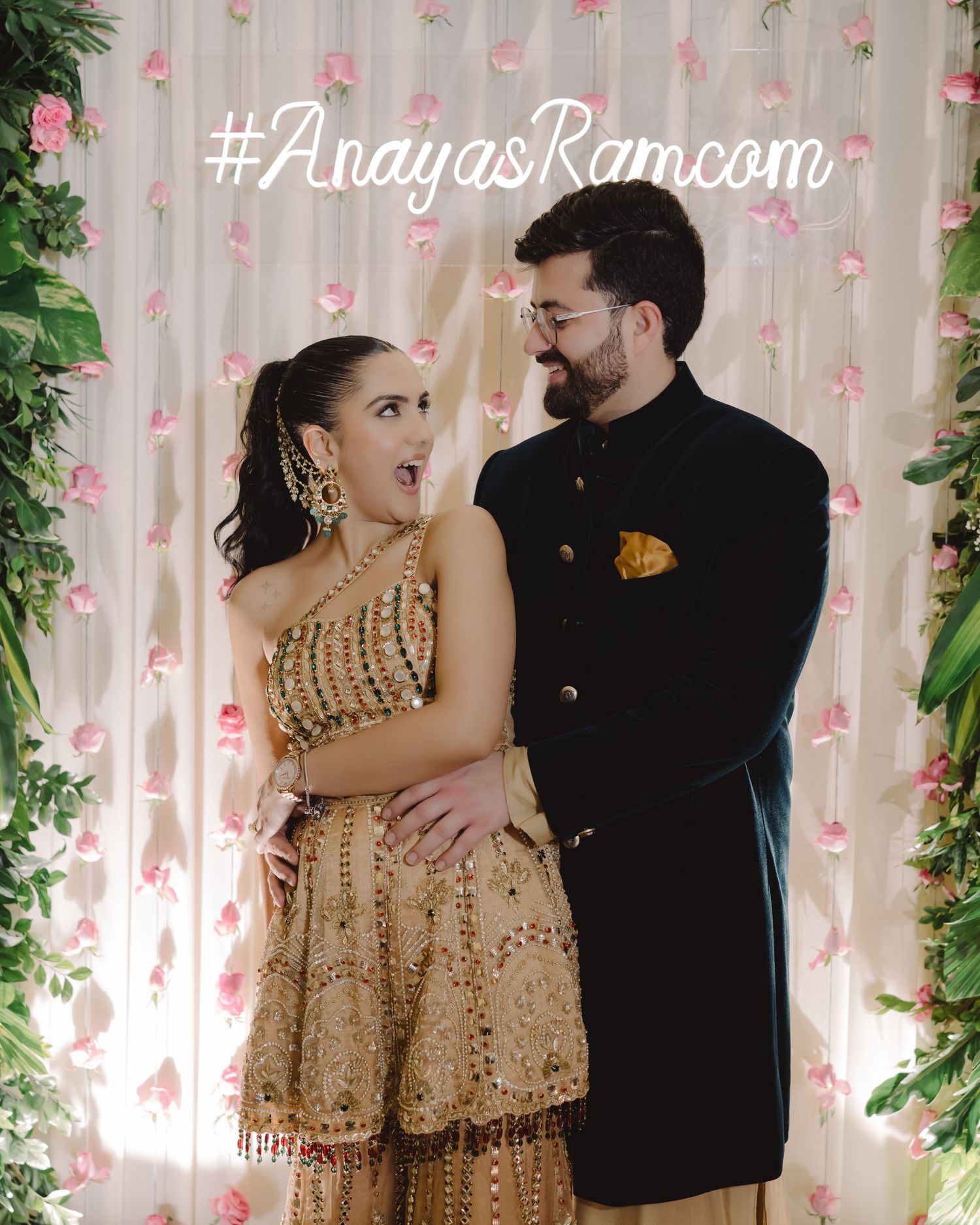 Couple in traditional Indian wedding attire posing joyfully before floral backdrop with hashtag AnayasRam.com.