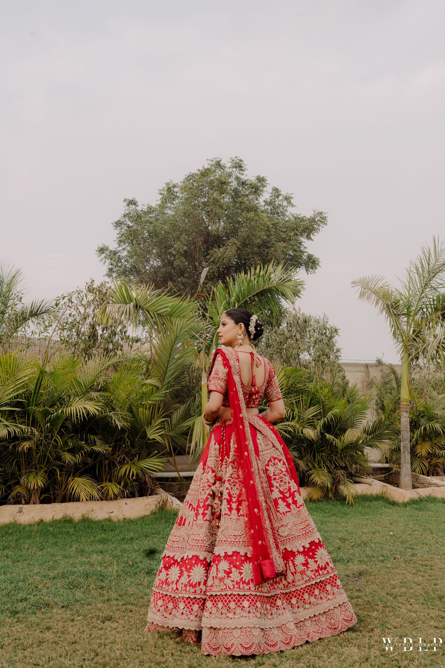 Bride in red embroidered lehenga choli standing in garden, showcasing intricate golden bridal embroidery and dupatta.