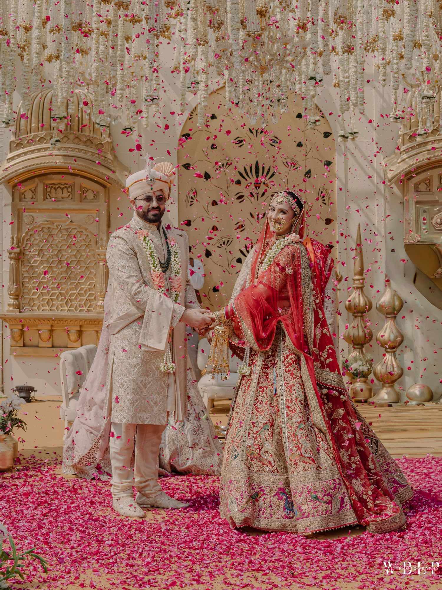 Indian bride in red lehenga and groom in ivory sherwani holding hands at floral wedding ceremony.