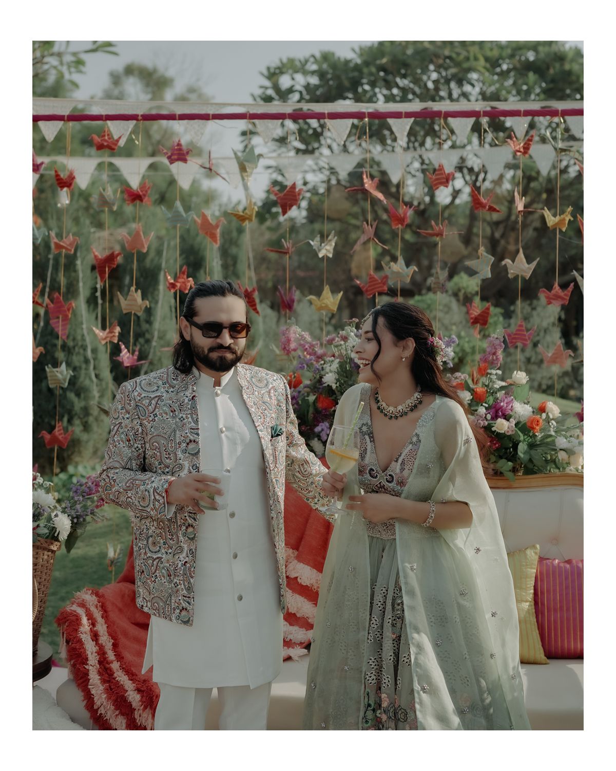 Couple stands beneath floral decor at outdoor celebration with hanging red and white flowers against greenery backdrop.