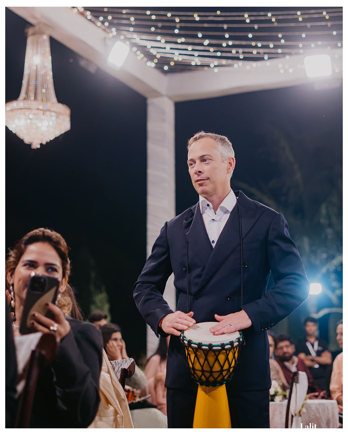 Guests enjoy a drum performance under string lights and a crystal chandelier at an outdoor reception.