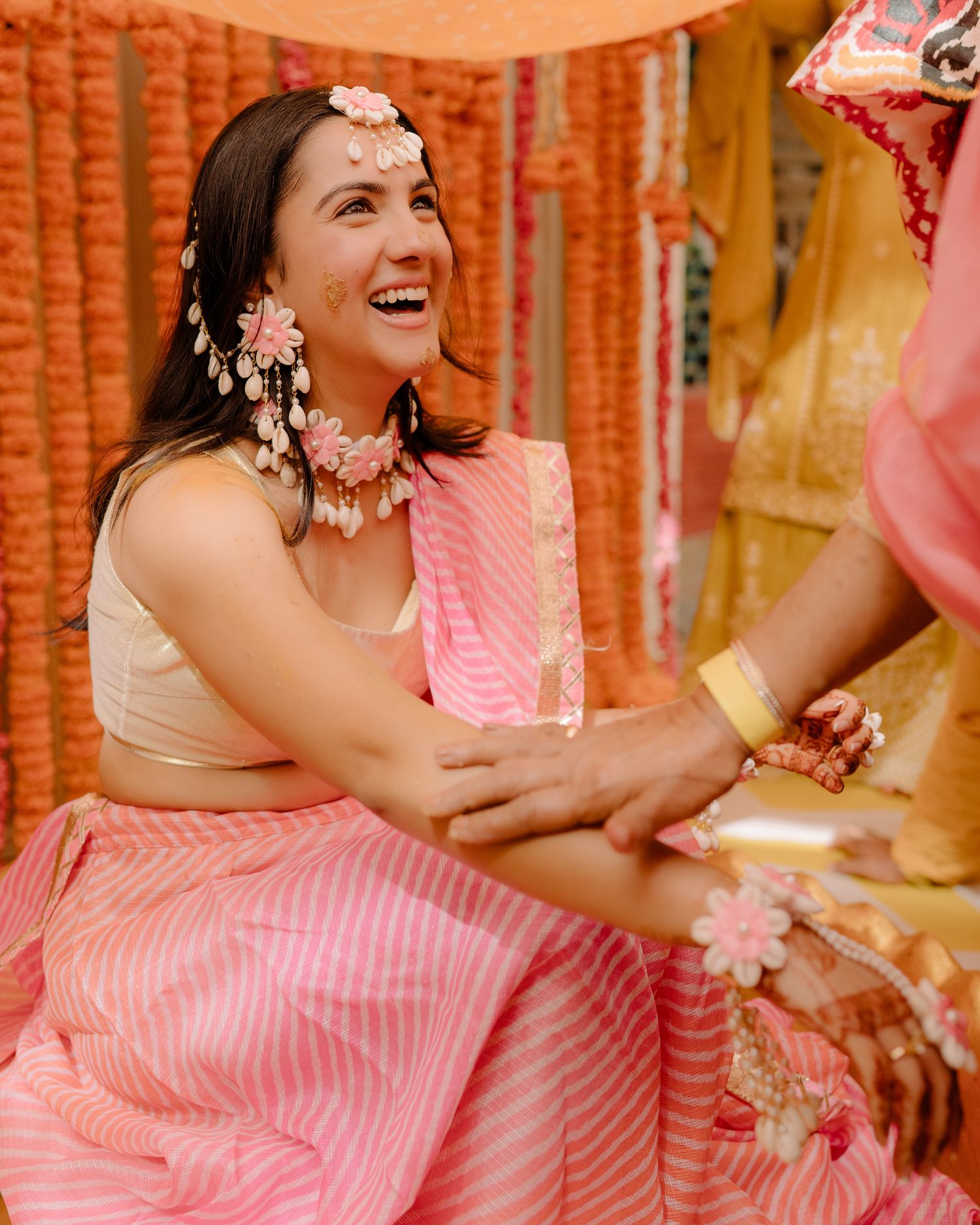 Bride laughing joyfully in pink lehenga with floral jewelry during Indian wedding mehndi ceremony.